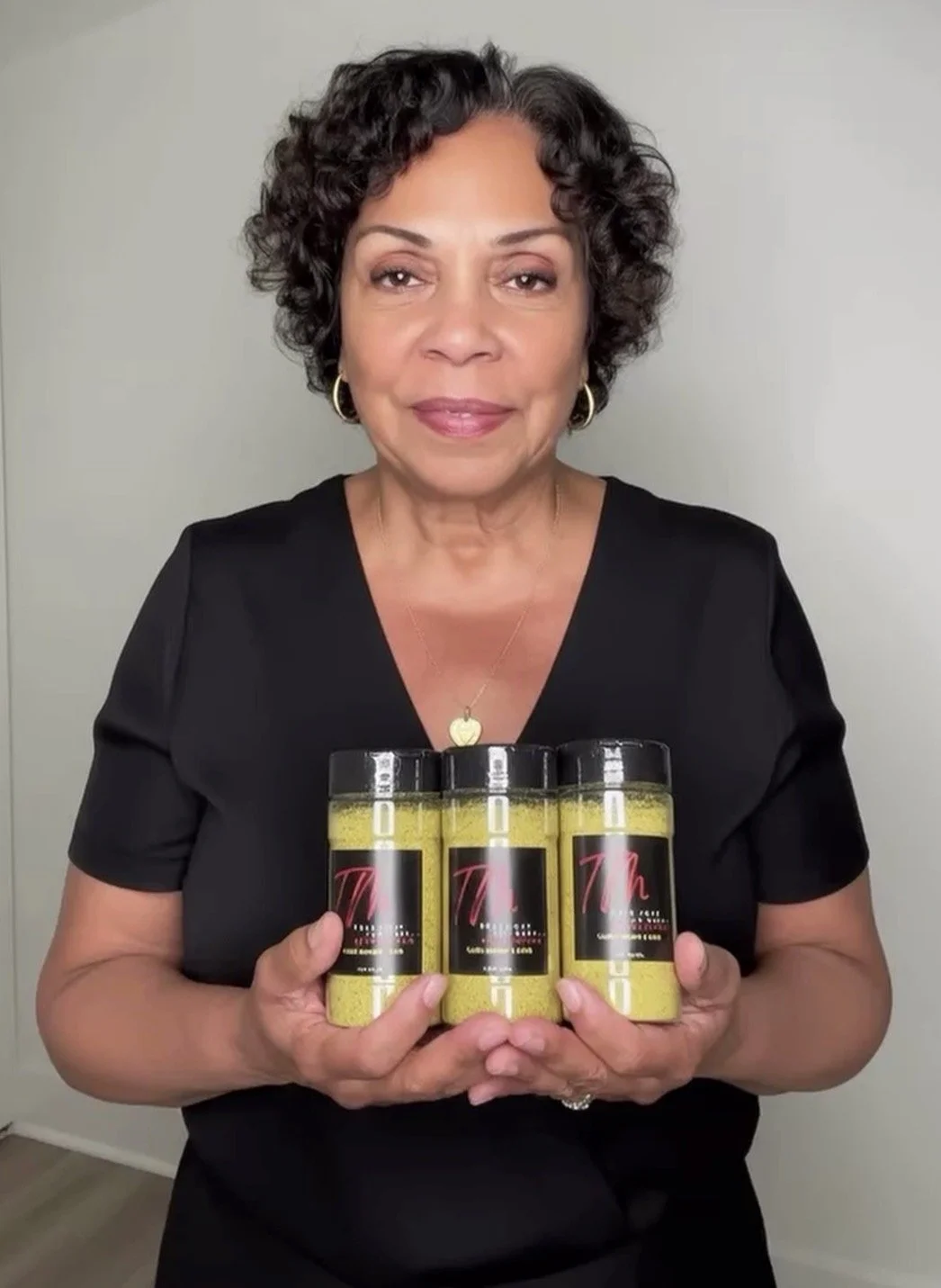 A woman with short curly hair wearing a black top is holding three containers of a yellow product and smiling at the camera.