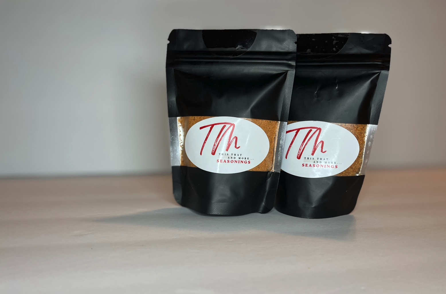 Two black resealable pouches of seasoning labeled 'This That and More... Seasonings' with a white oval and red and black text, placed on a light-colored surface.