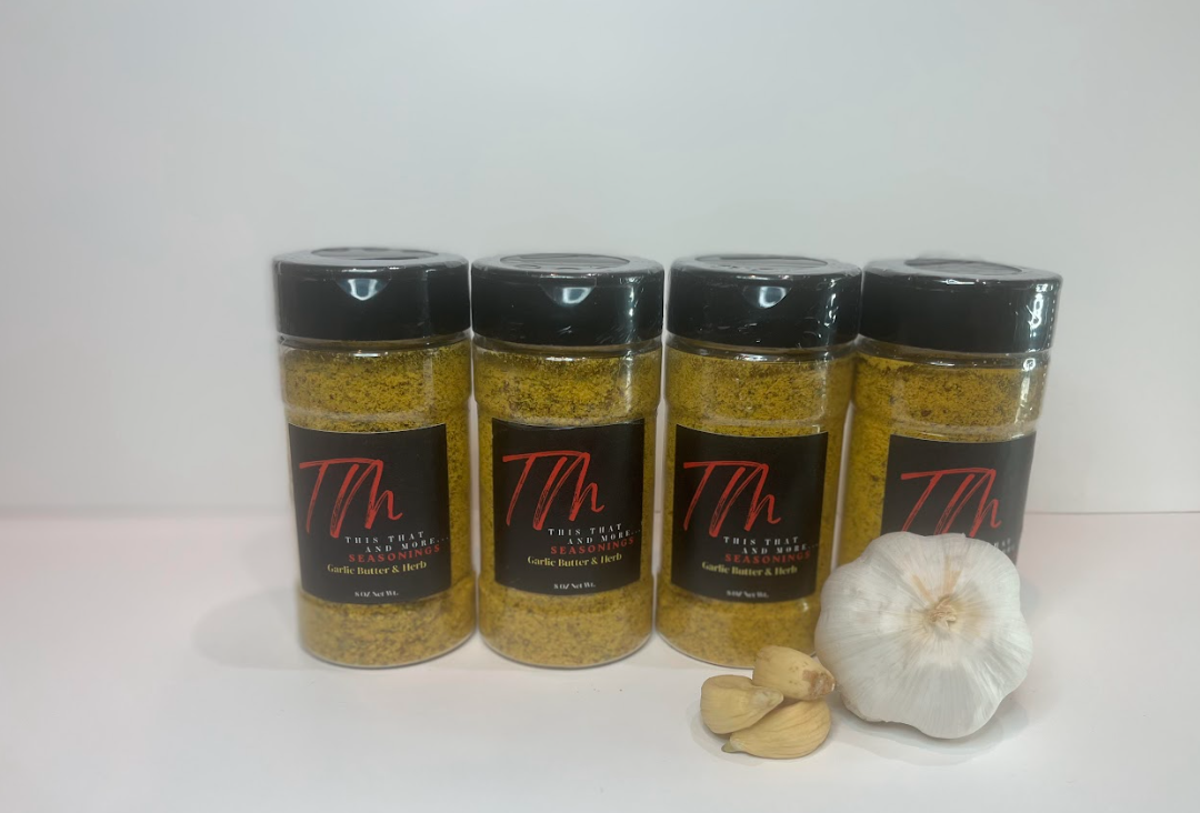 Four bottles of seasoning labeled 'Garlic Butter & Herb' placed in a row, with a head of garlic and two garlic cloves in front of them.