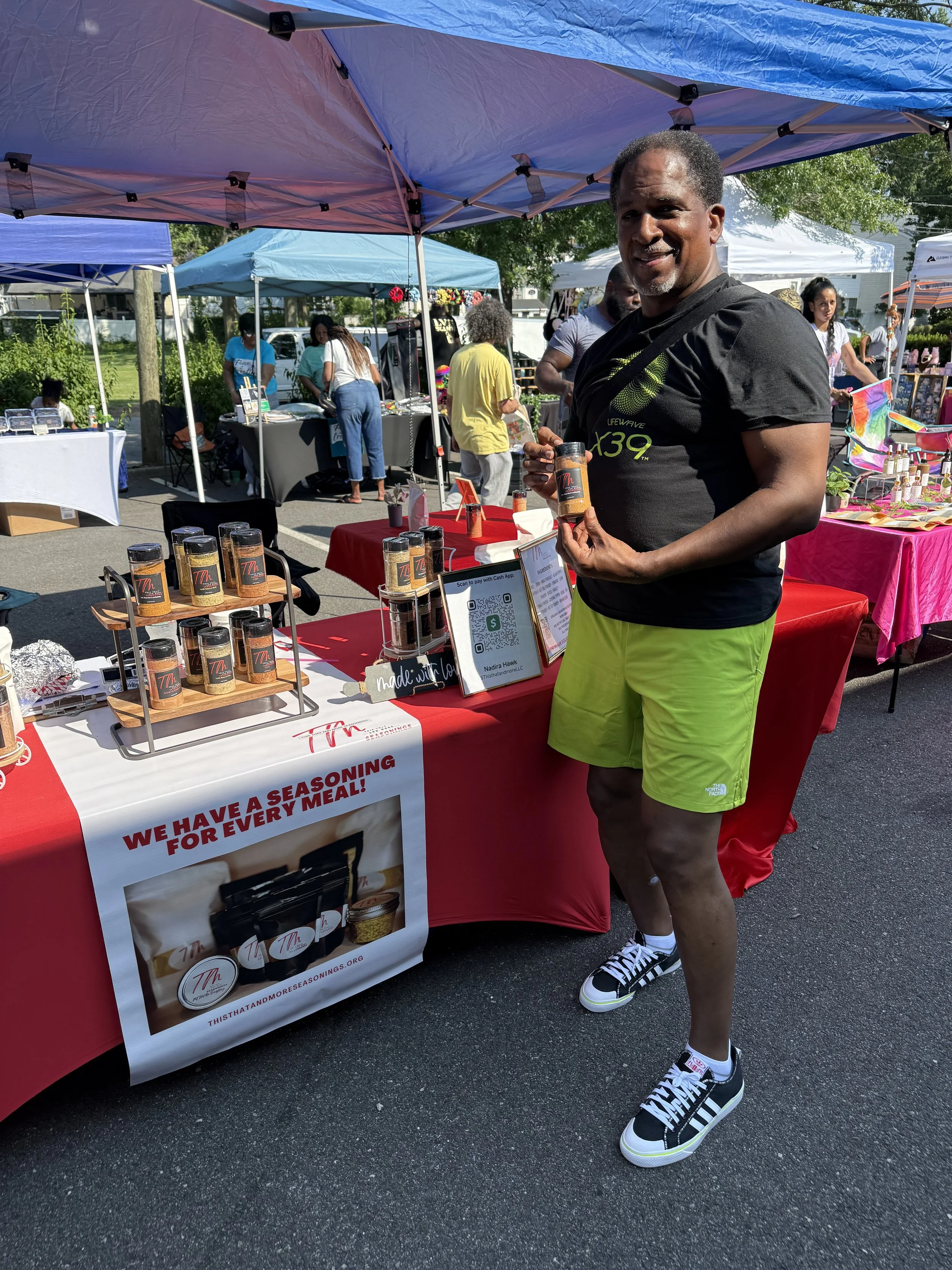 A man at an outdoor marketplace holding a jar of seasoning, standing next to a booth with other jars of seasoning and a sign that reads, 'We have a seasoning for every meal!'