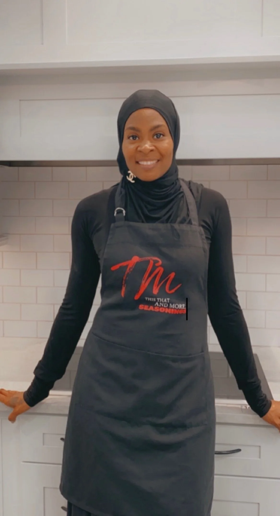 Smiling woman in black headscarf and apron standing in a kitchen with white tile backsplash.