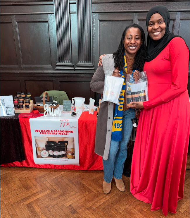 Two women smiling and posing together in front of a display table with seasonings, spice jars, and promotional items. One woman is wearing a gray coat and the other is wearing a red dress and a black headscarf. They are holding small bags of seasonin