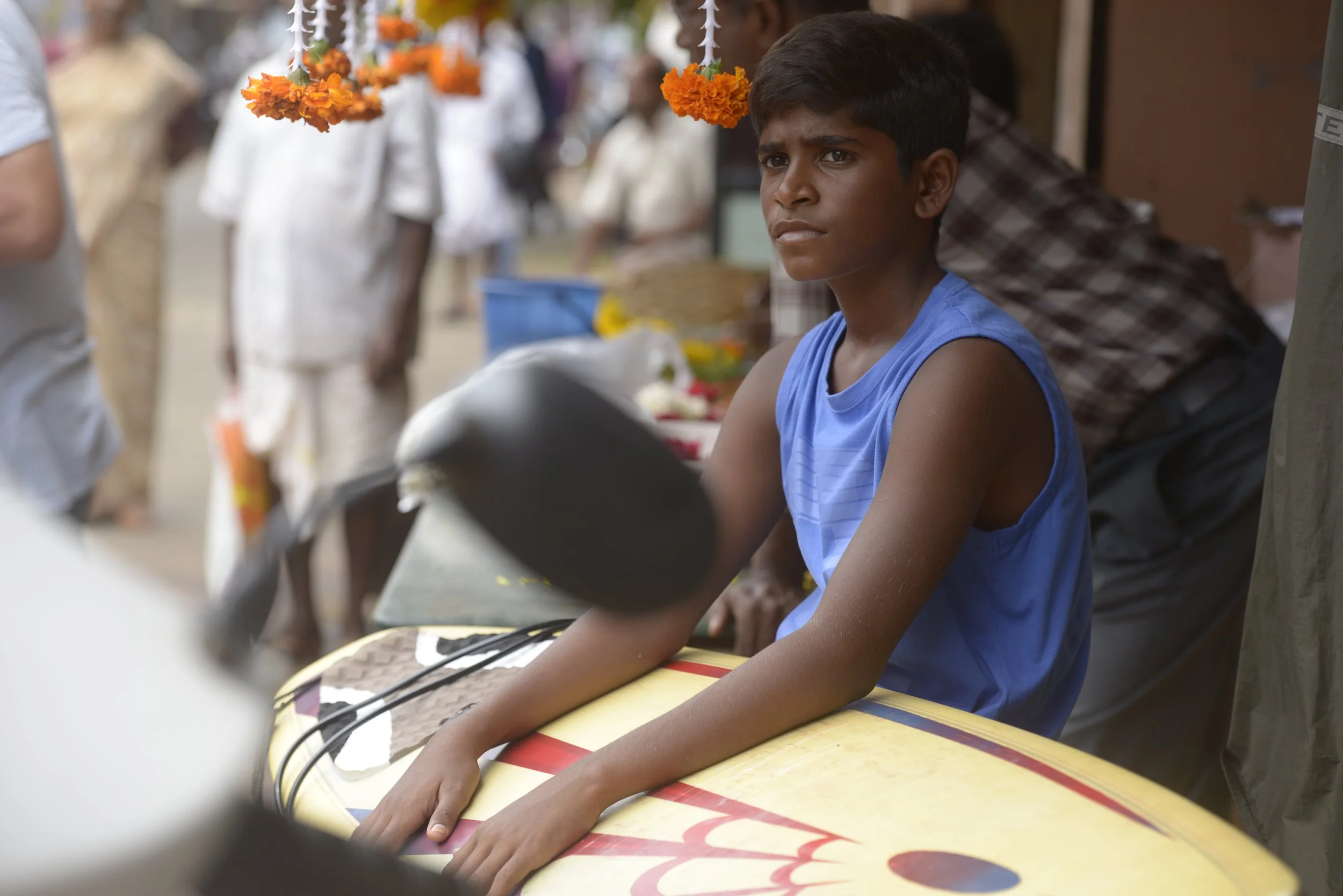 A young boy sitting at a table in a busy outdoor marketplace, looking thoughtful or concerned, with a surfboard in front of him.