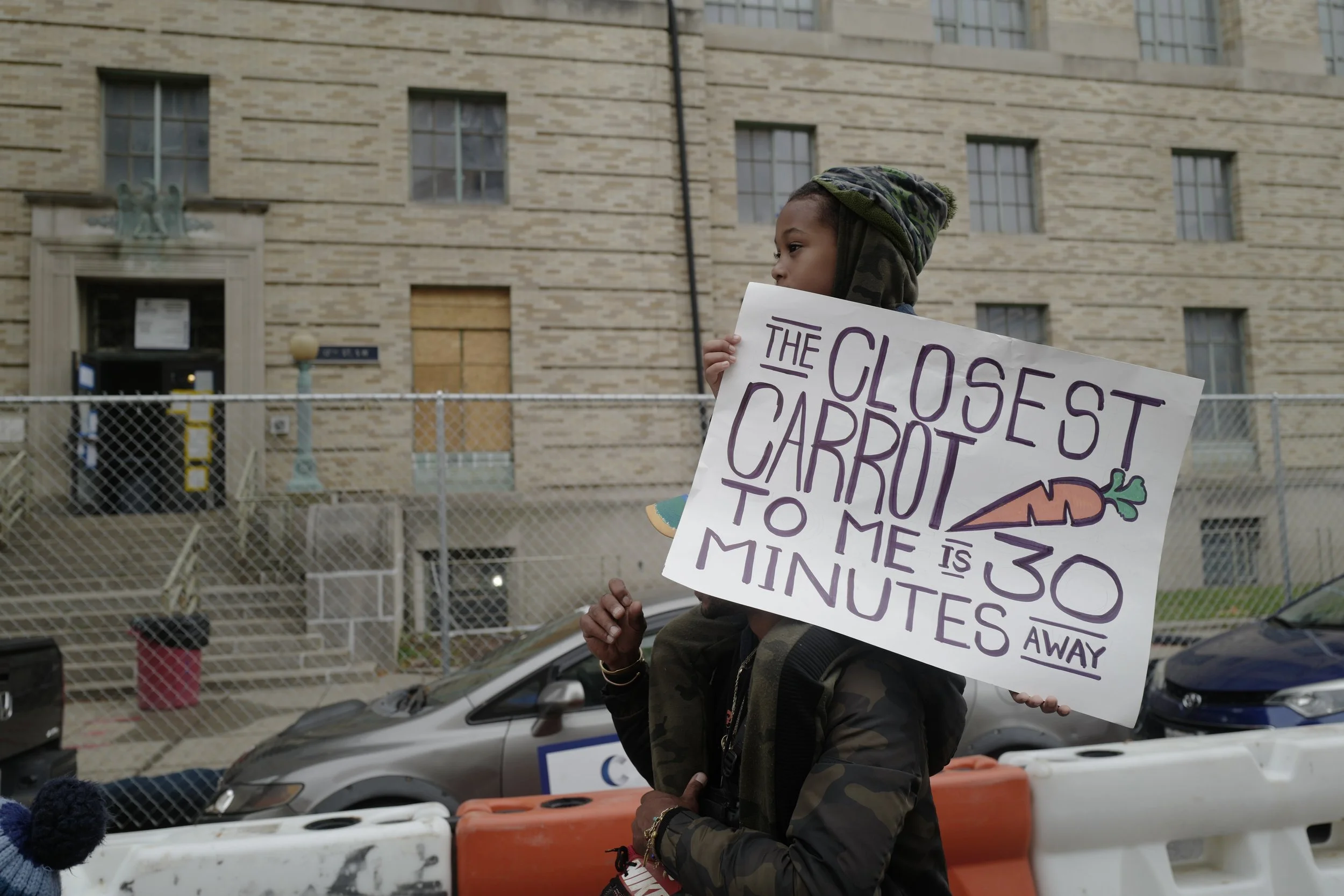 A young person wearing a hat and jacket holding a protest sign that reads 'The closest carrot to me is 30 minutes away' with a drawing of a carrot, standing near construction barriers and parked cars in front of a building.