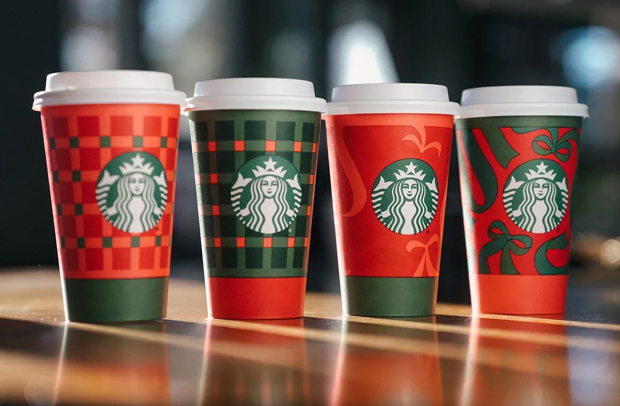 Four Starbucks hot beverage cups with holiday-themed designs, arranged in a row on a wooden surface, with sunlight and blurred greenery in the background.