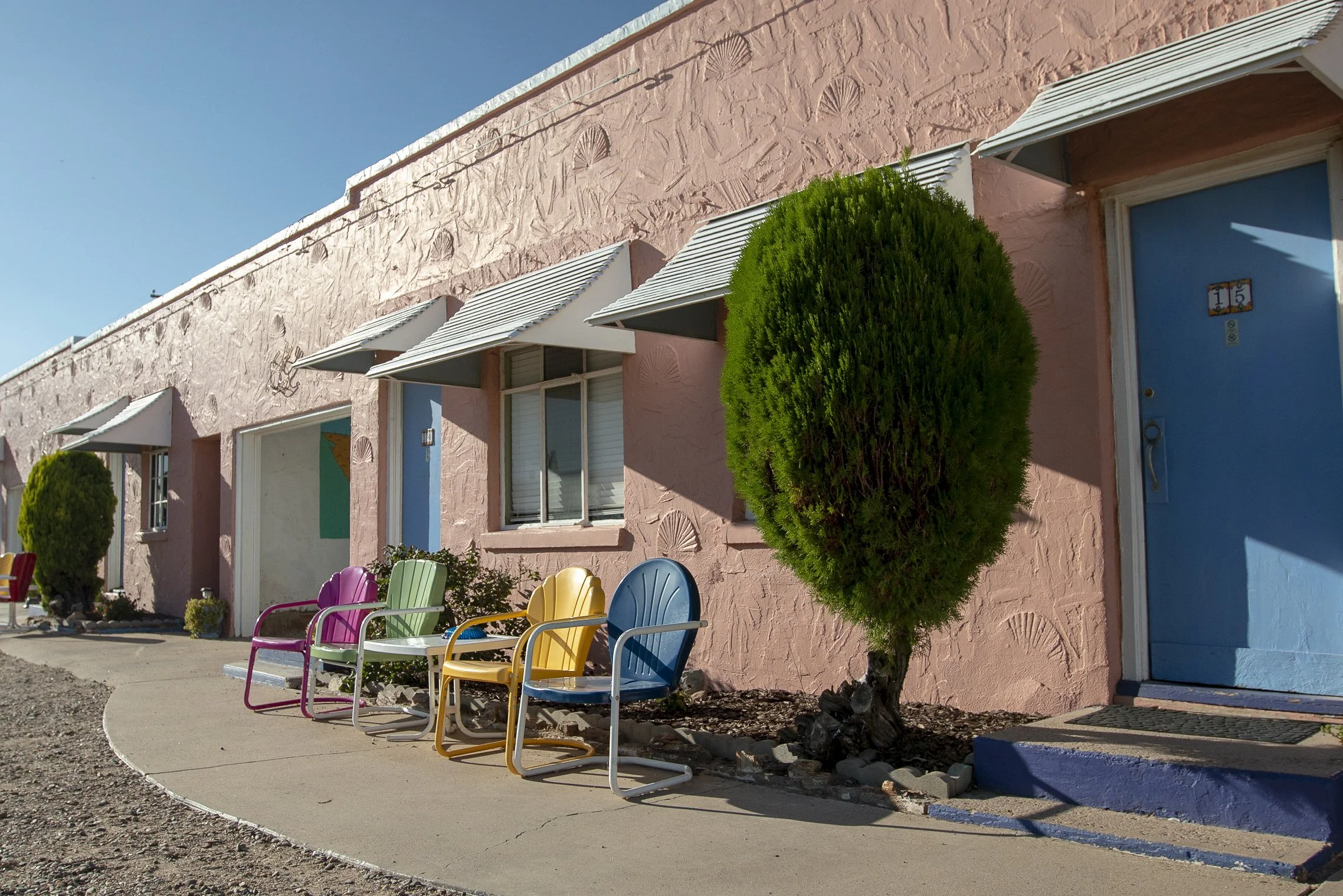 Pink building with blue doors and white awnings, colorful chairs outside, and a manicured green tree in front.
