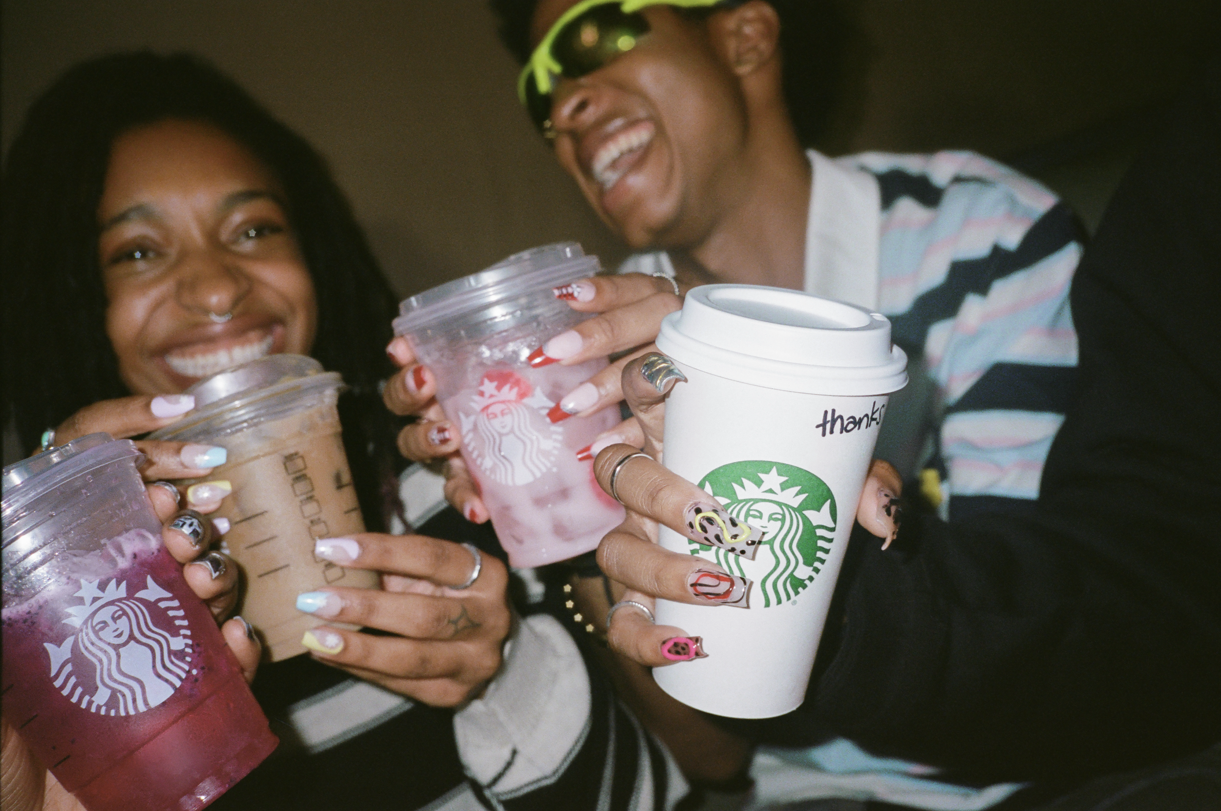 Two women smiling and holding Starbucks drinks, one with a pink drink, the other with a coffee, and a man smiling holding a white Starbucks cup with 'thanks' written on it.