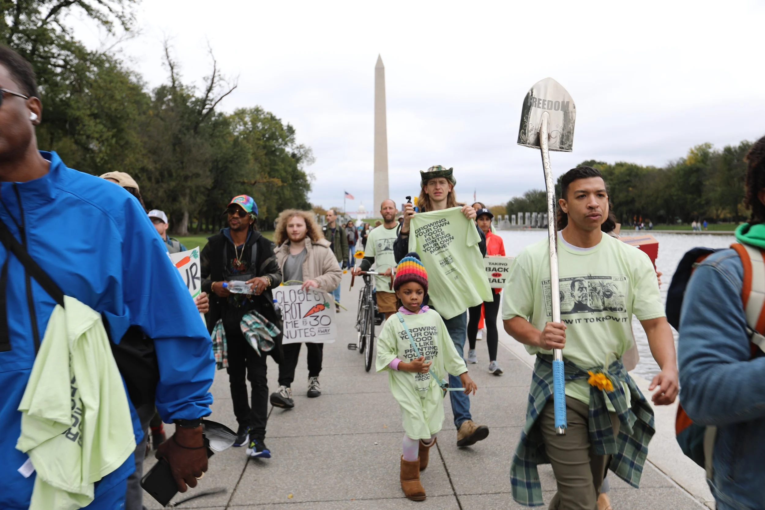 Group of diverse people participating in outdoor protest or march near the Washington Monument, holding signs and banners about social or political issues on cloudy day.