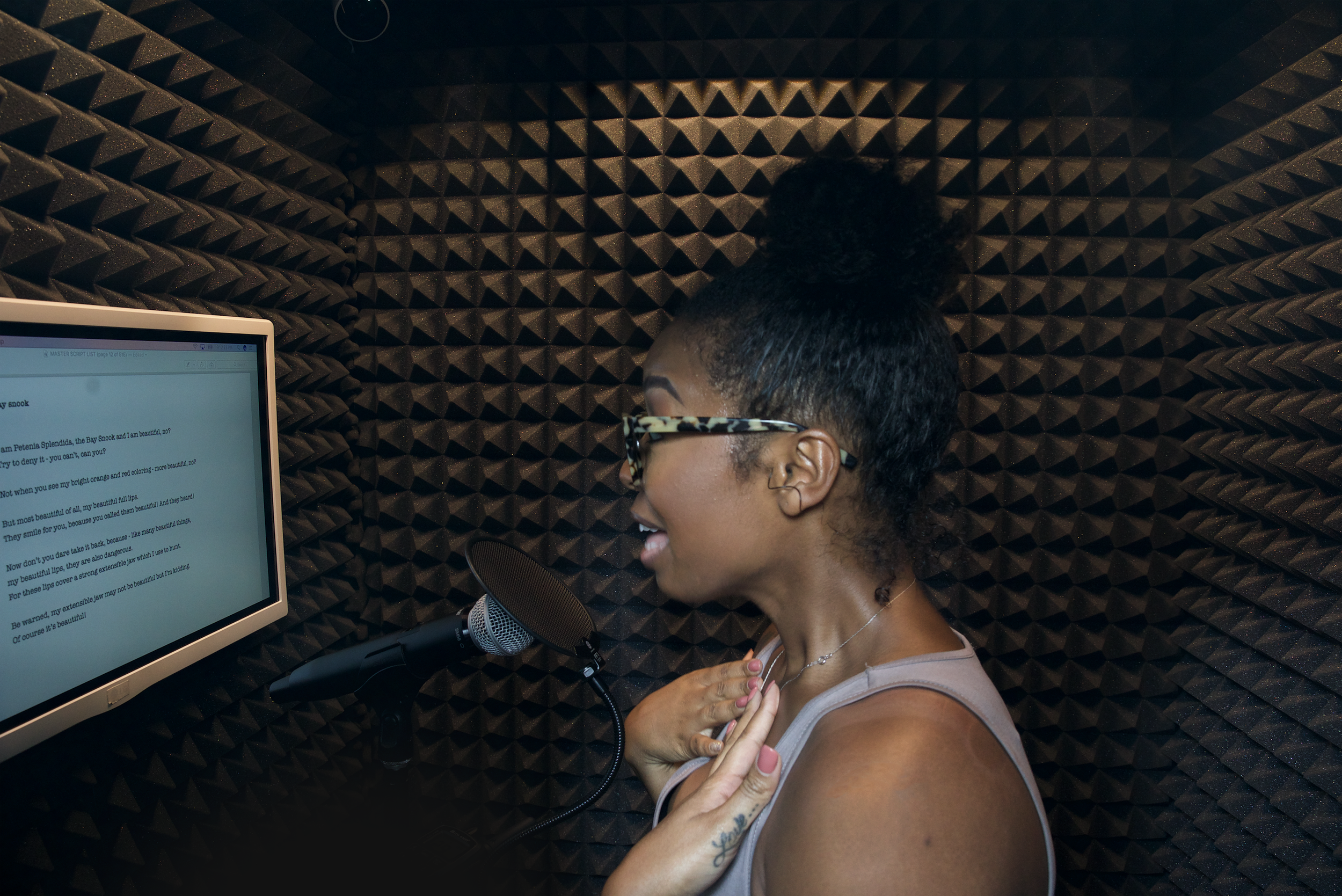 A woman singing into a microphone in a soundproof recording booth, wearing glasses and a sleeveless top.