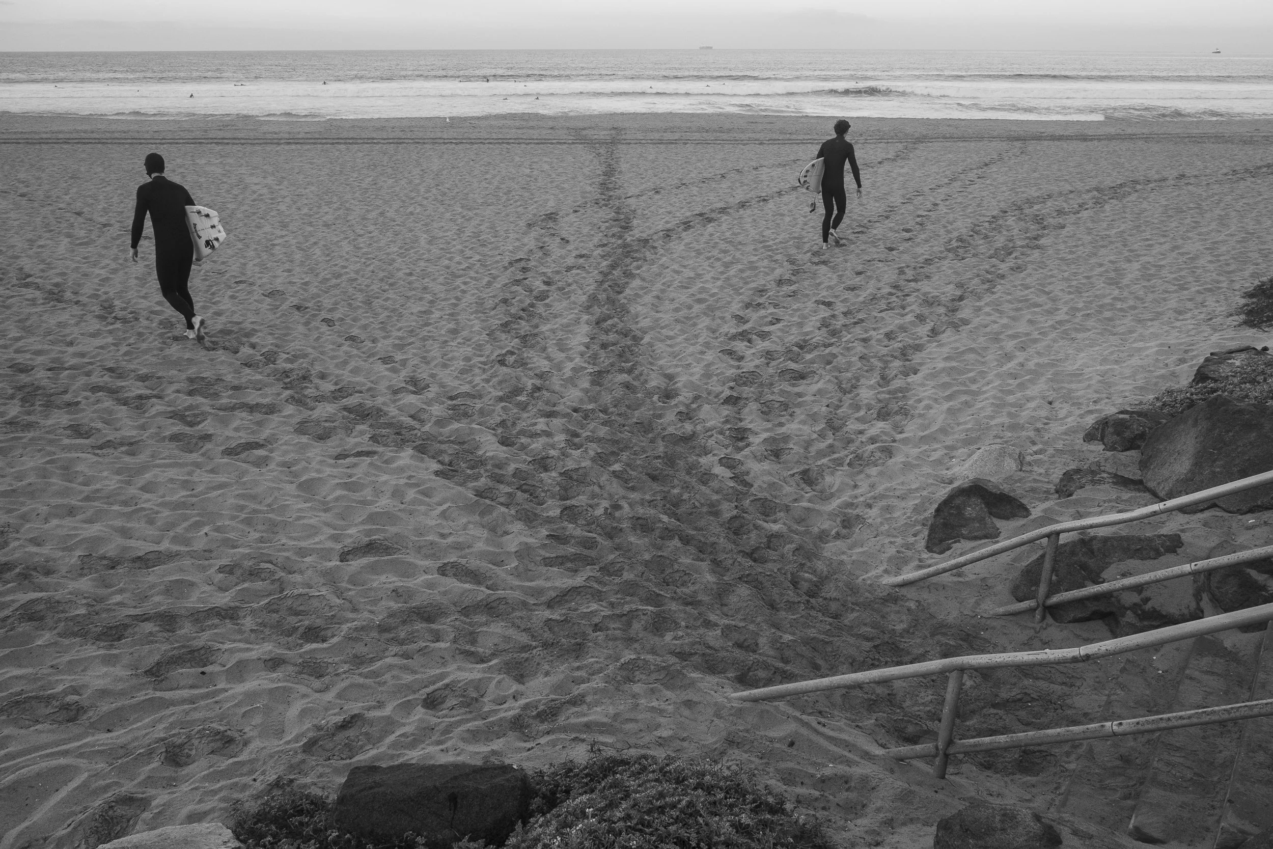 Two surfers carrying surfboards walk across sandy beach toward the ocean with gentle waves, black and white photo.