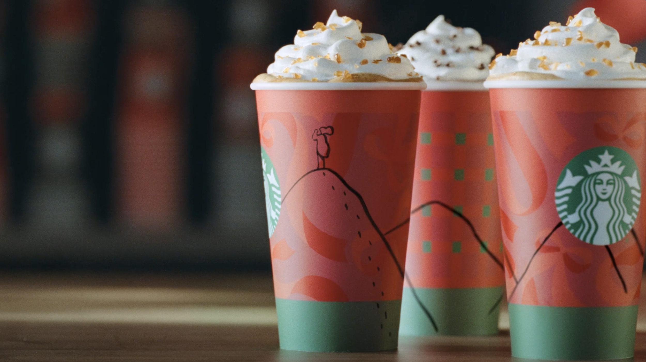 Three Starbucks holiday-themed cups filled with whipped cream and caramel topping, set on a wooden surface.