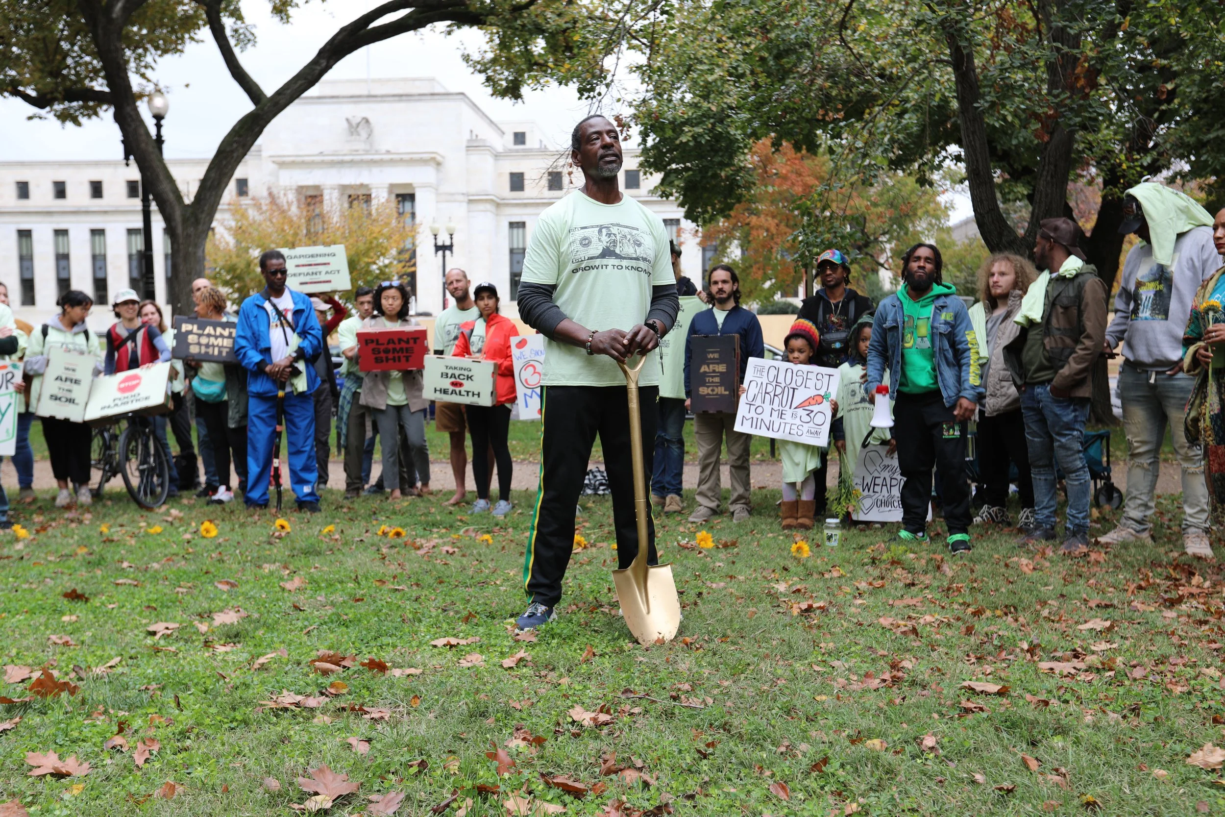 Group of people participating in a protest rally in a park, holding signs advocating for environmental issues, with a man in the foreground holding a shovel.