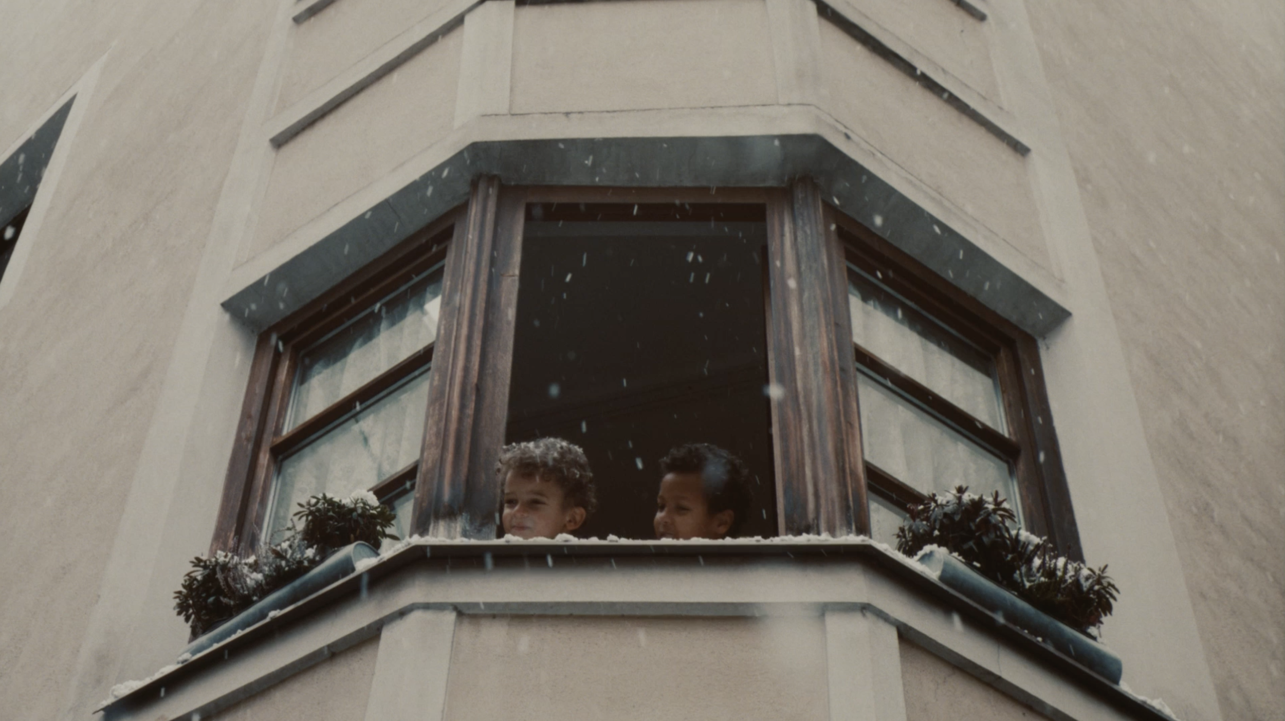 Two children looking out of a corner window of a building during a snowstorm. The window has wooden frames, and there are small potted plants on the windowsill covered with snow.