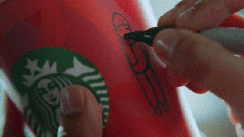 A person is drawing a cartoon character on a red Starbucks cup with a black marker, with the Starbucks logo visible on the cup.