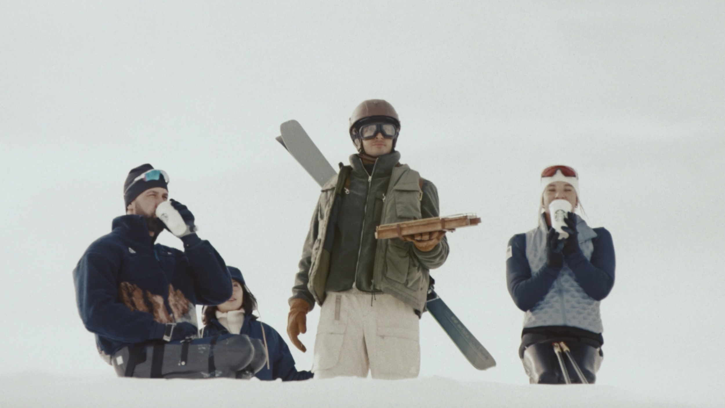 Four people in winter clothing, standing on snow, with some holding drinks, and winter sports gear such as helmets, goggles, and snowboards.