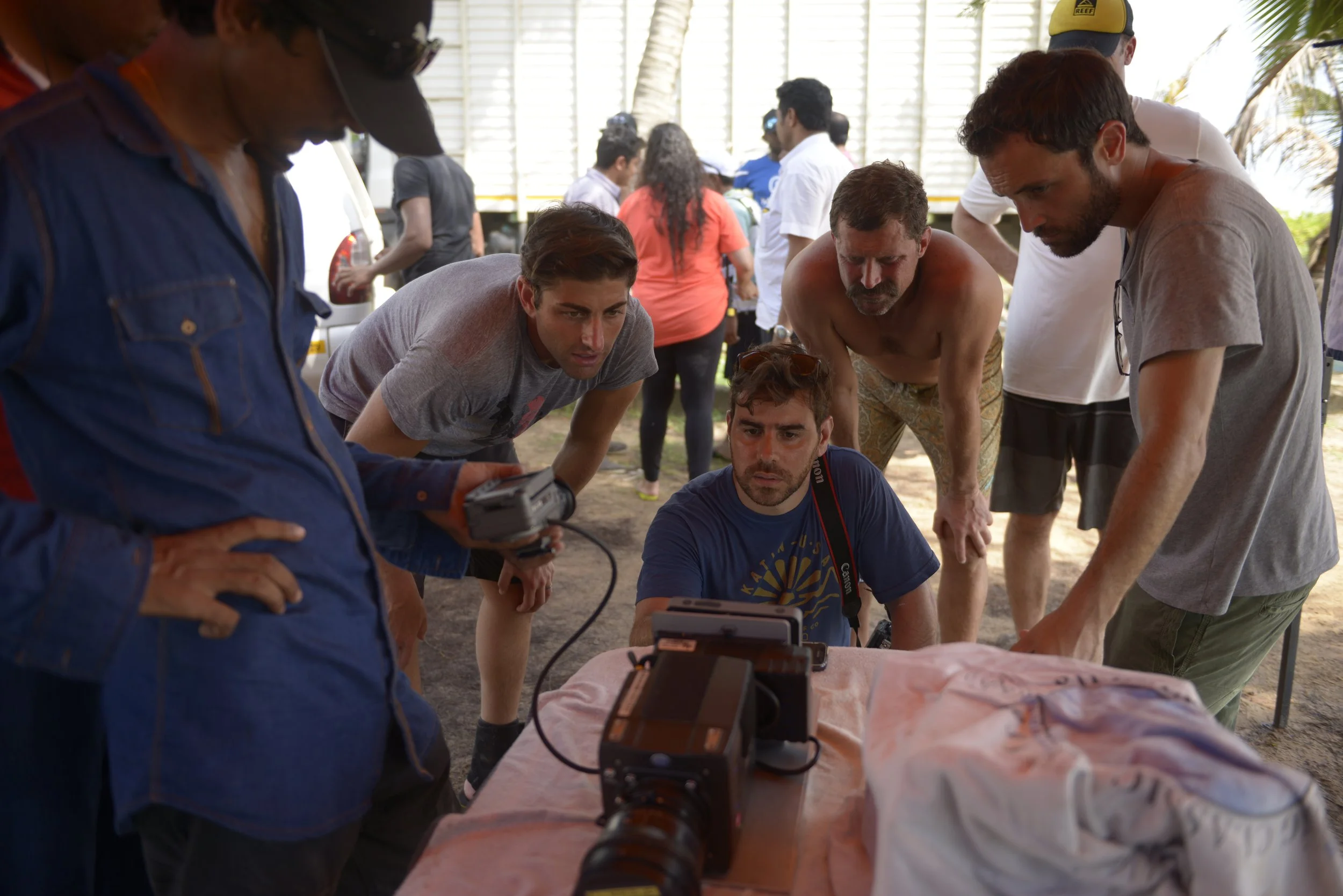 Group of people gathered around a camera and equipment, watching attentively at an outdoor event, with some looking serious and focused.