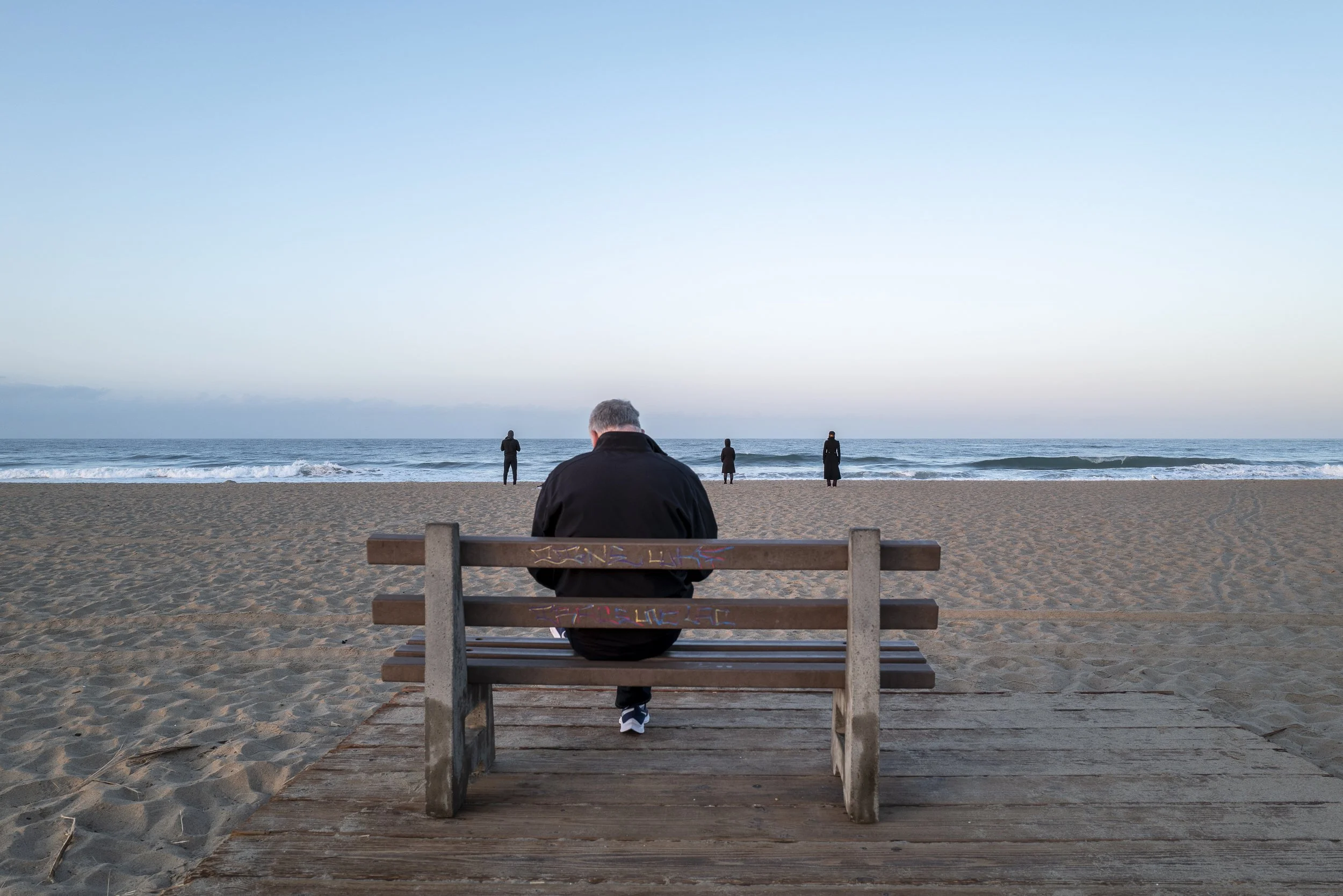 A person sitting on a wooden bench on the beach facing the ocean, with three people standing near the water in the distance, and a clear sky above.