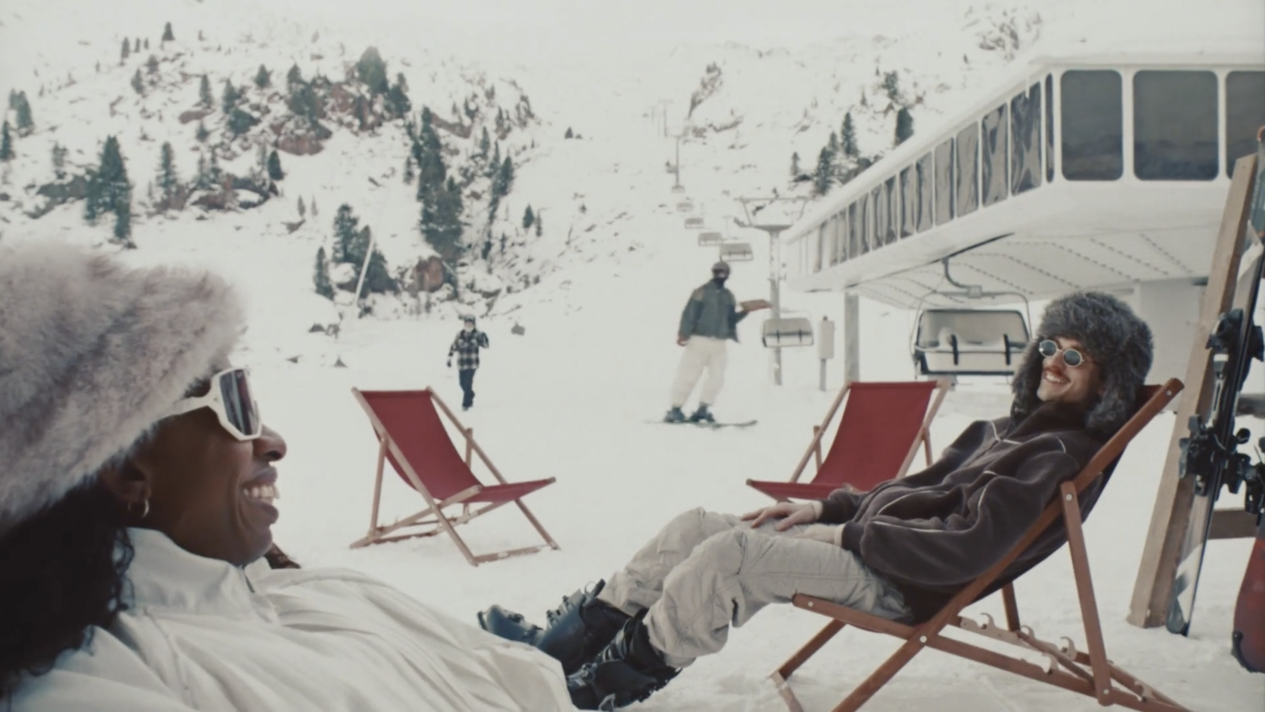Two people sitting outdoors in snow at a ski resort, smiling and relaxing in lounge chairs, with skiers and snowy mountains in the background.