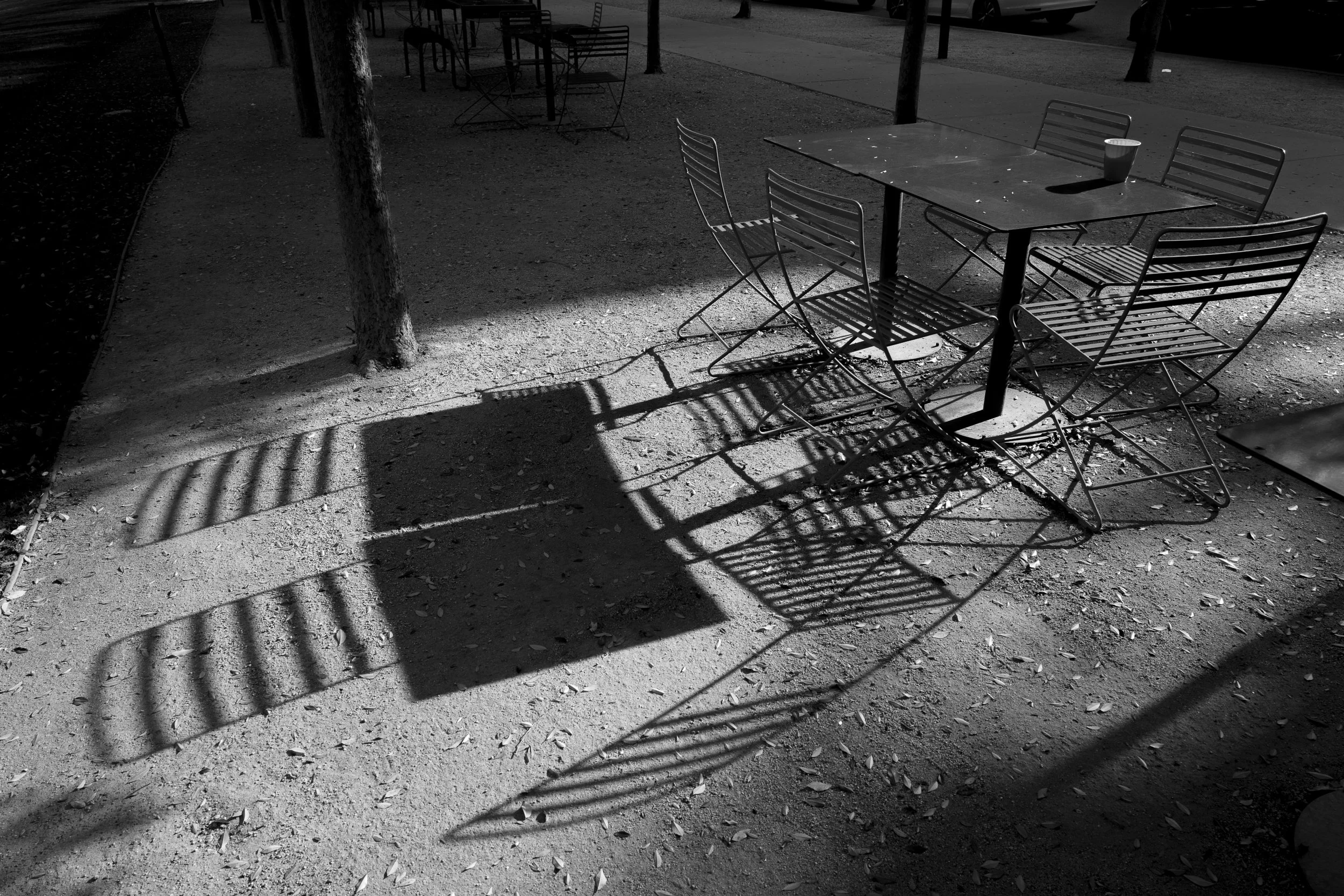 An outdoor patio area with a table and four chairs. The chairs cast shadows on the ground, along with the shadow of a nearby tree and a rectangular object, possibly a bench or a crate. The scene is in black and white.