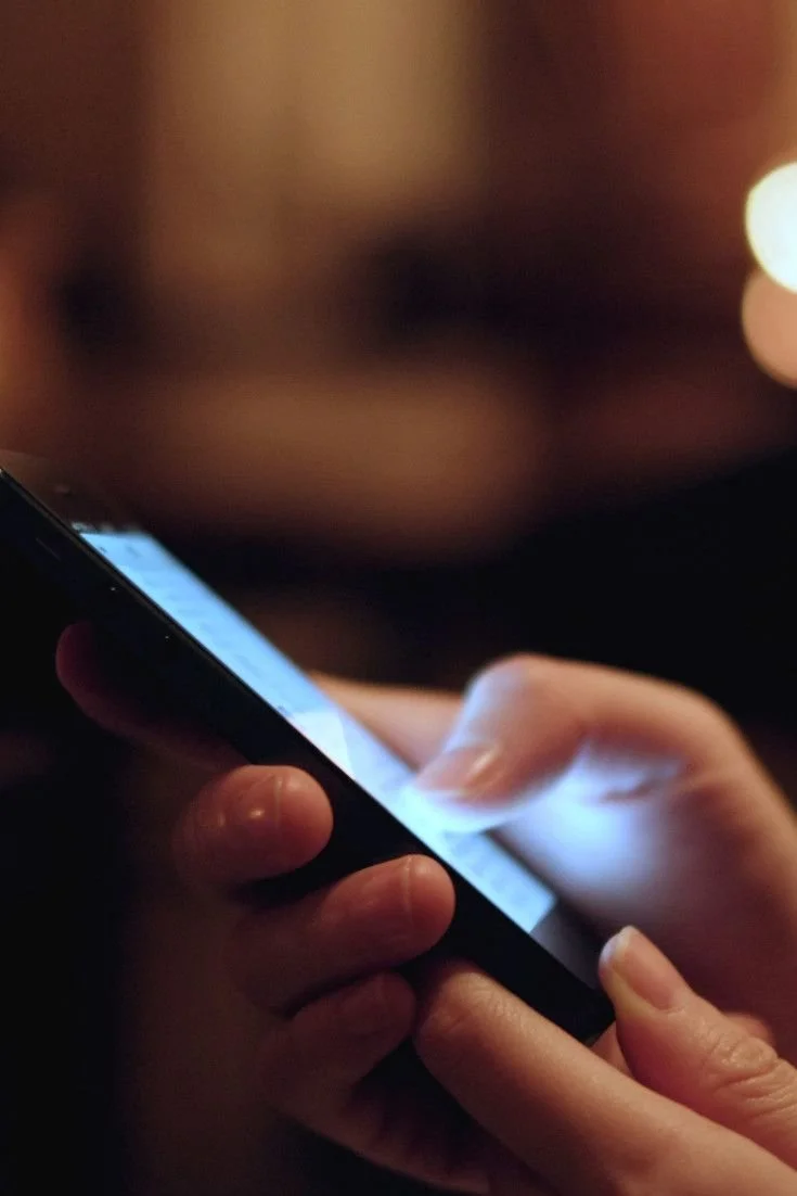 A person holding a smartphone with both hands, with the screen lit up and fingers touching the screen, in a dimly lit environment.