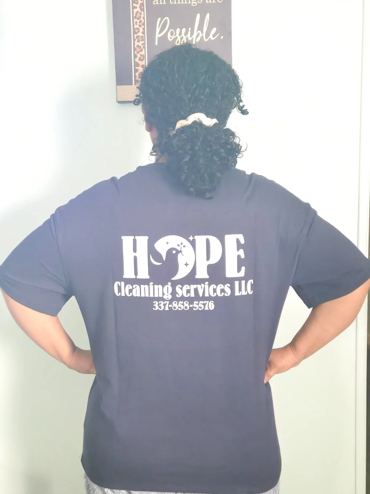 Back view of a person with curly hair tied back with a hair tie, wearing a navy T-shirt with white text and graphics on the back, standing in front of a white wall with a motivational poster.