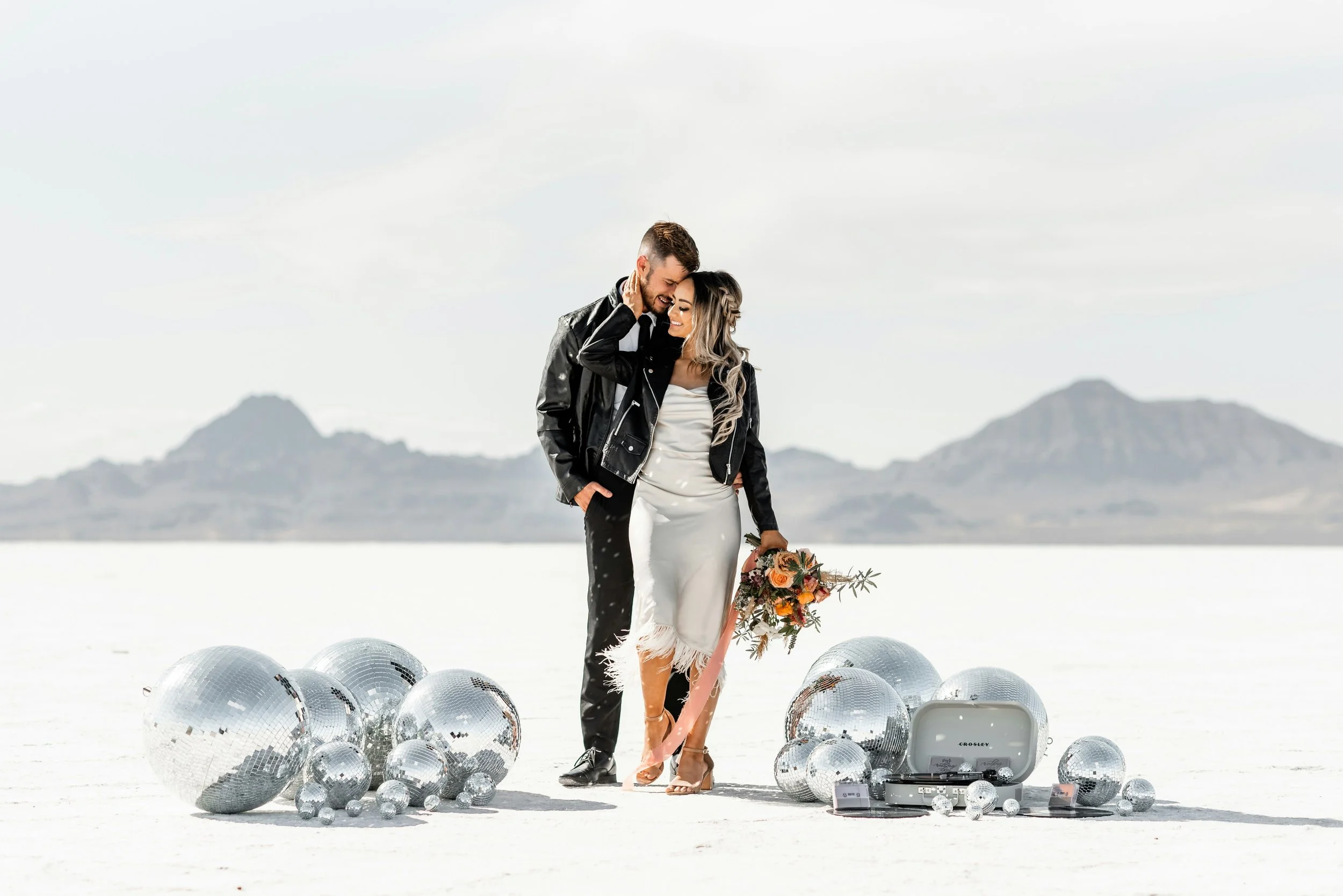A couple dressed in wedding attire standing on a reflective white surface, surrounded by disco balls, with a mountain range in the background. The woman is holding a bouquet of flowers, and they appear happy and close together.
