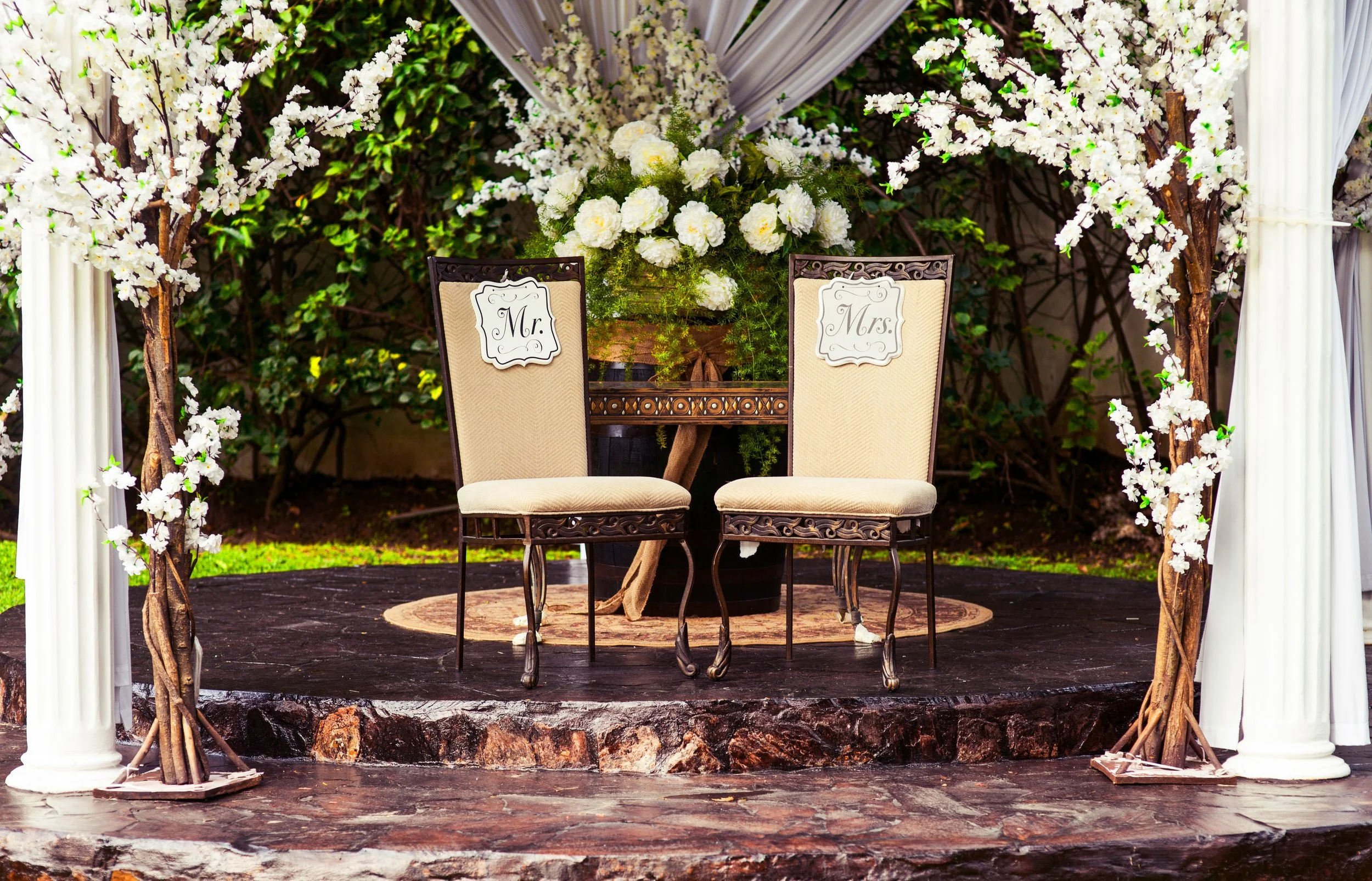 A decorated wedding altar with two beige chairs labeled 'Mr.' and 'Mrs.', surrounded by white flowers and greenery.