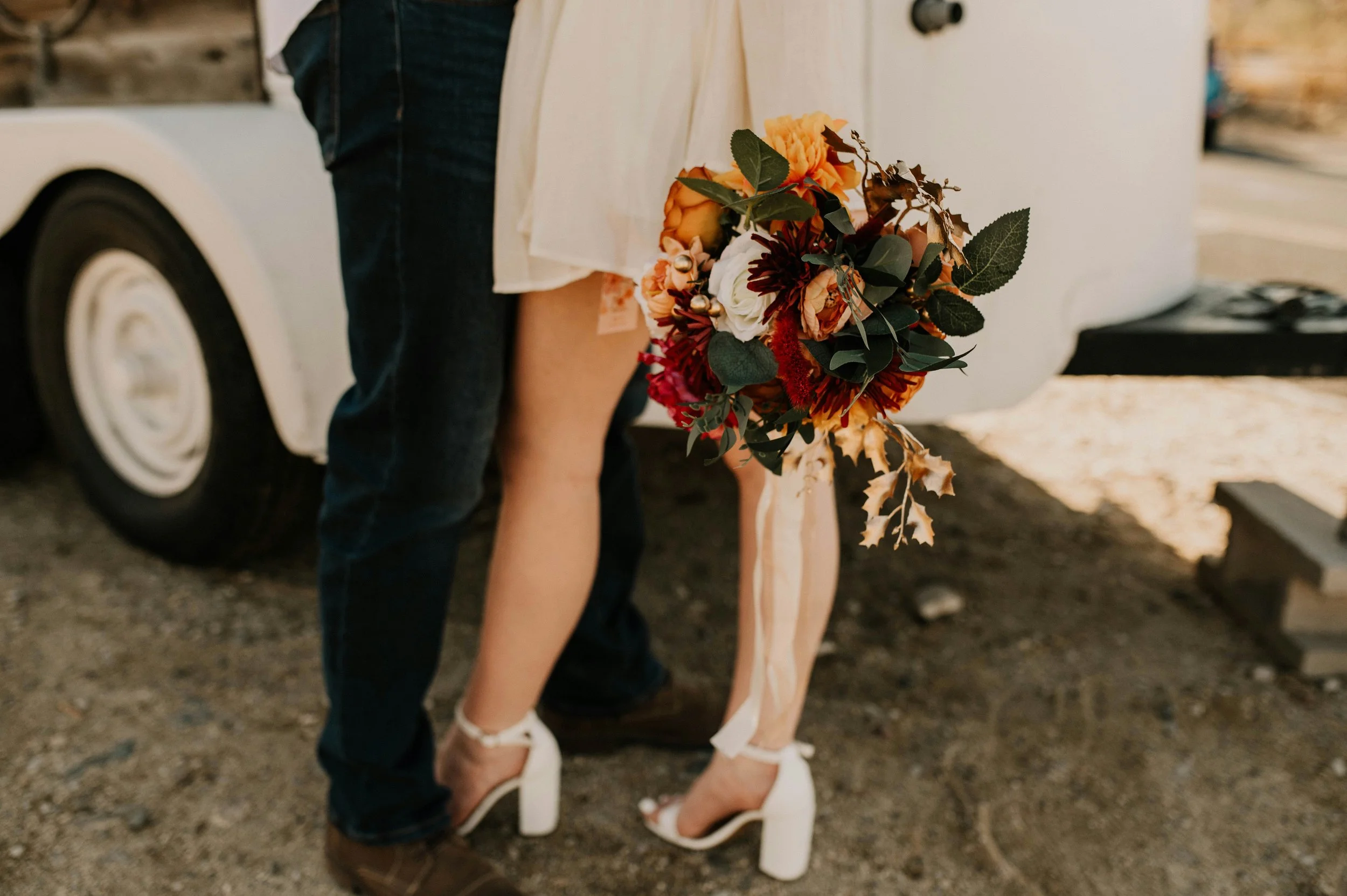 A person holding a bouquet of flowers standing near a vintage white vehicle on a gravel surface.