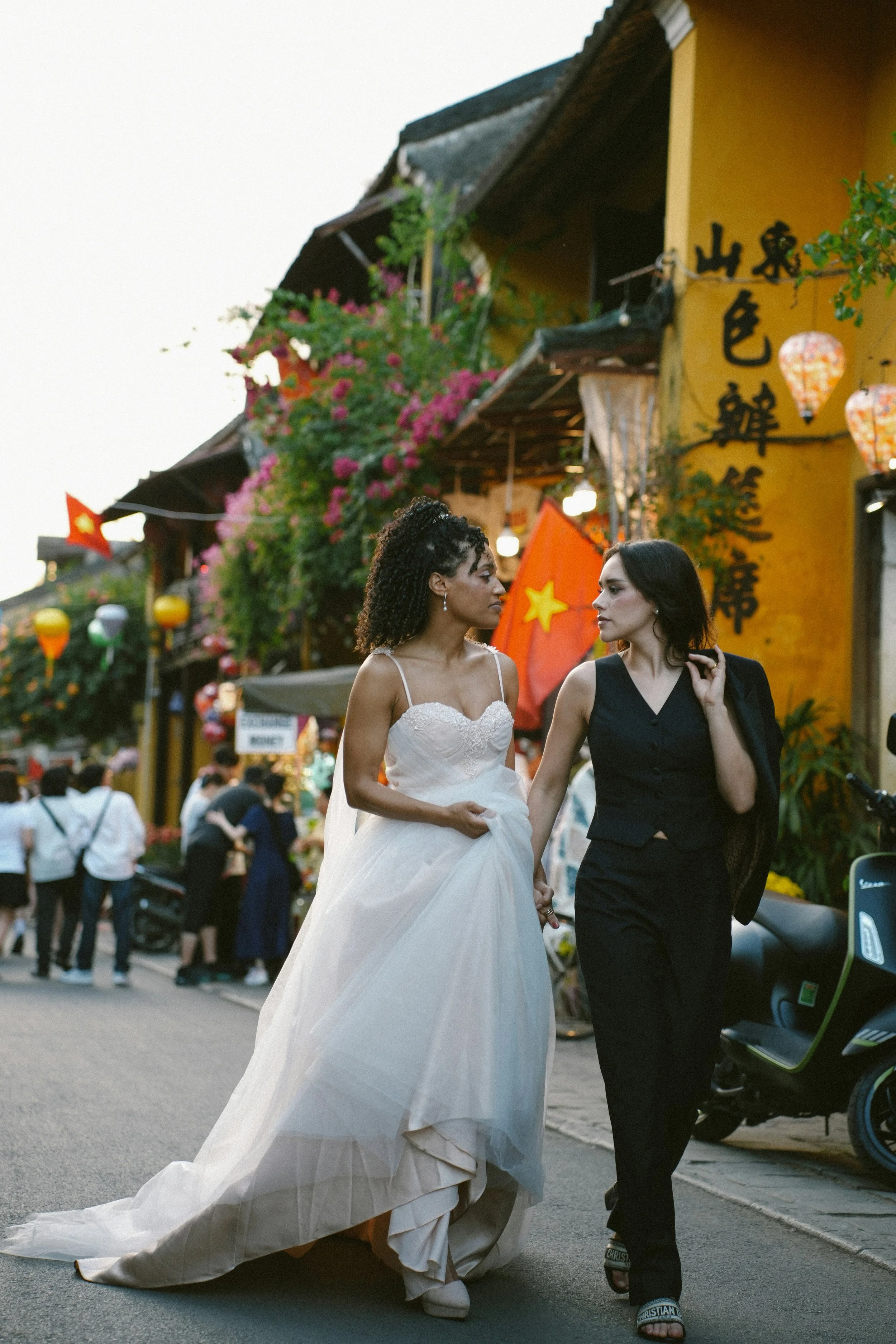 Two women walk arm in arm on a lively street decorated with colorful lanterns and flowers. One woman wears a white wedding dress, and the other is in a black suit with a blazer draped over her shoulders.