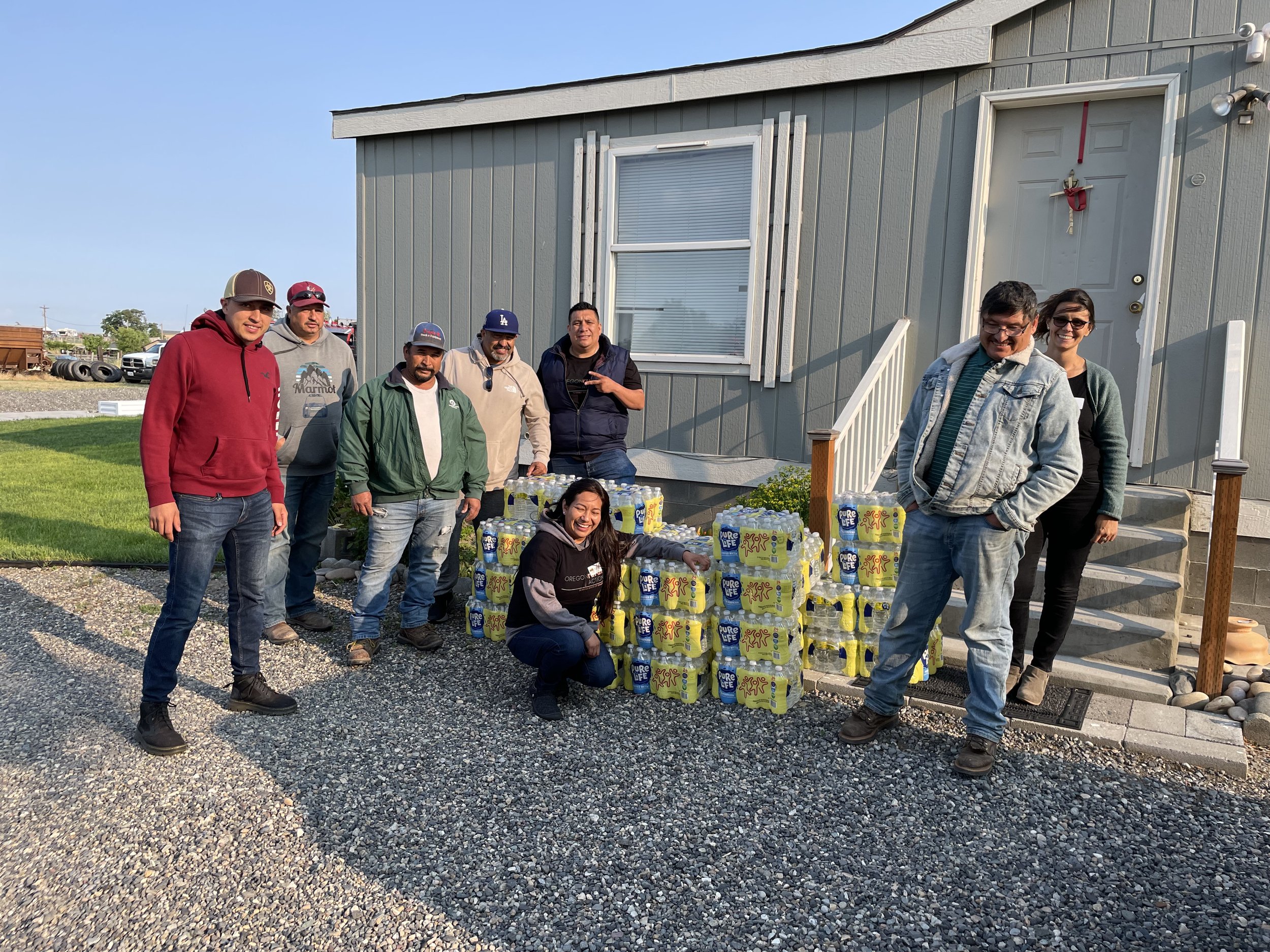 Group of people standing outside near a table filled with bottled drinks in front of a gray house with a front door and window, during daytime.