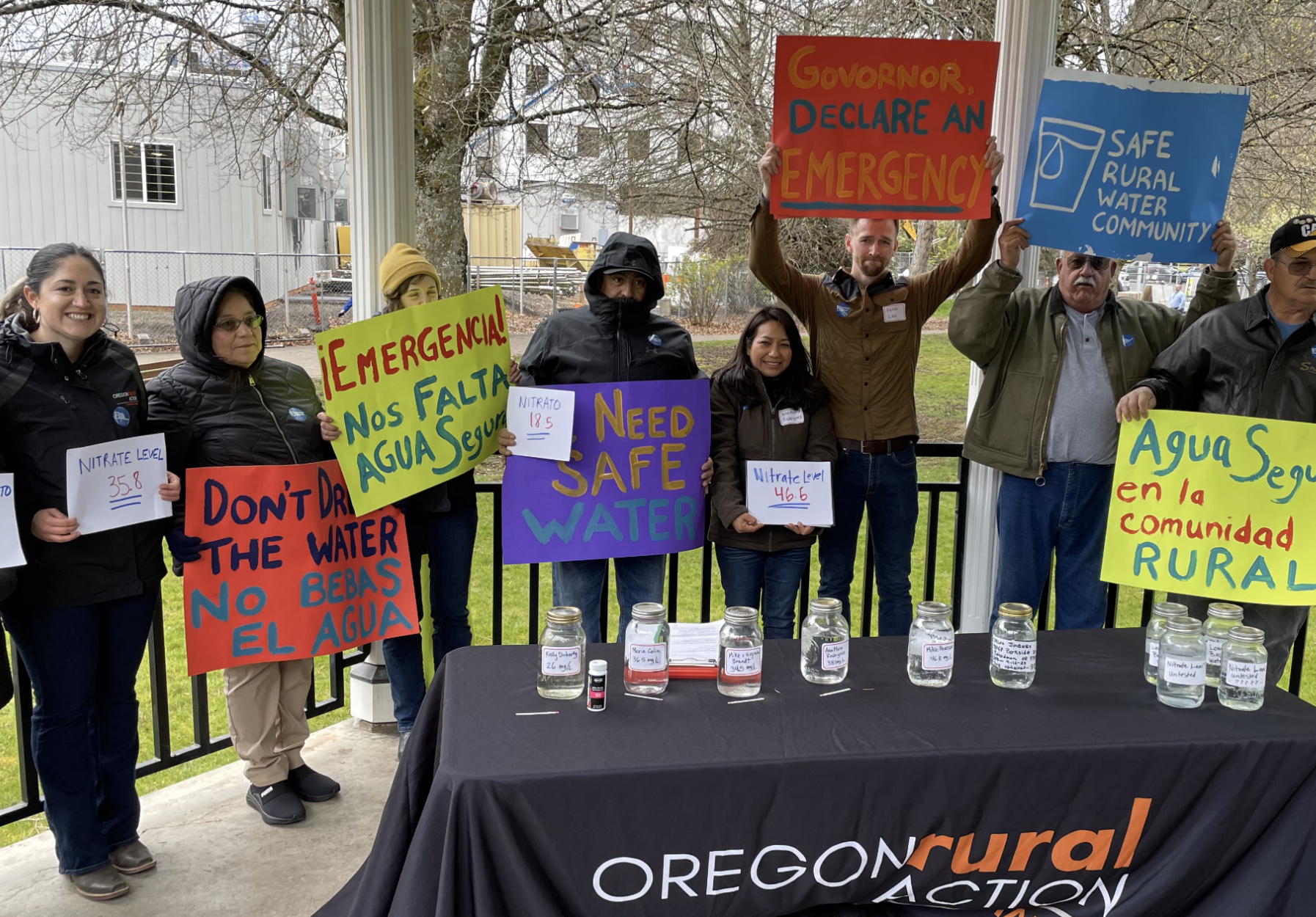 A group of people holding signs advocating for clean water and environmental issues at a rally or protest. Signs include messages about water safety, emergency, and rural community water access. There are water jars on a table in front of them.