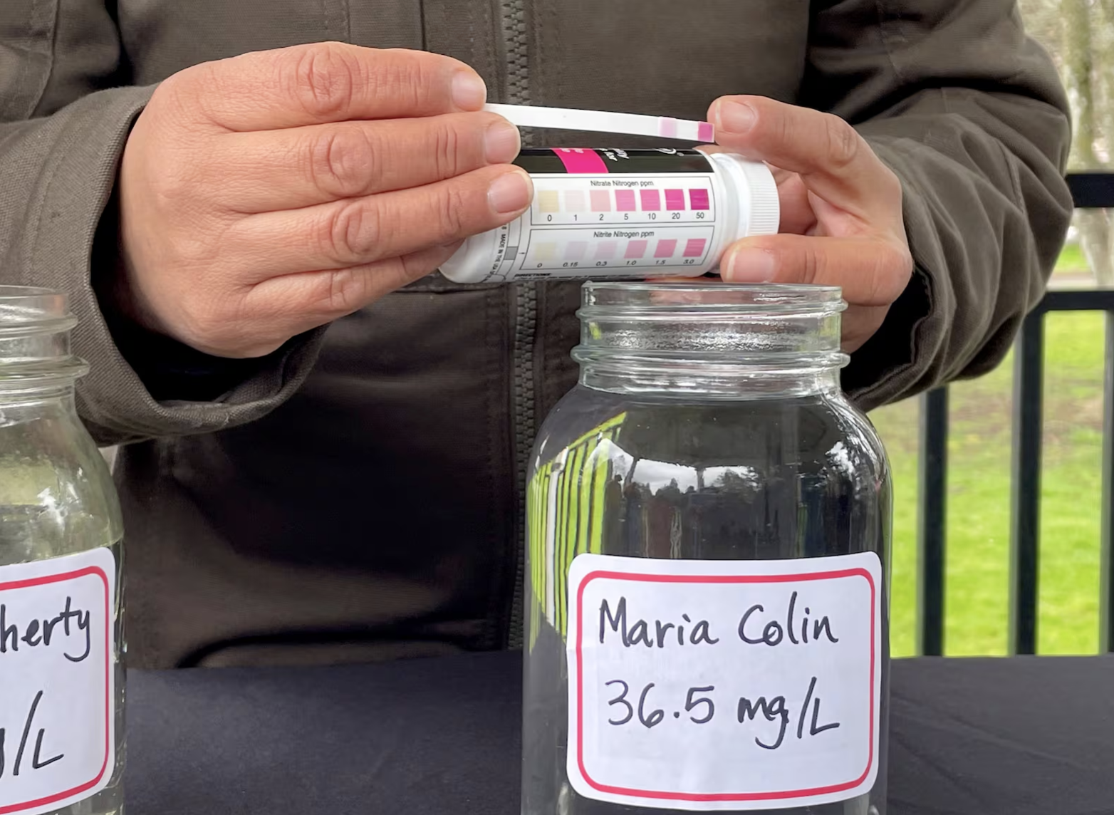 Person holding a urine test strip over a jar labeled with a name and medical information, with another jar visible beside it. The test strip shows a pink line.