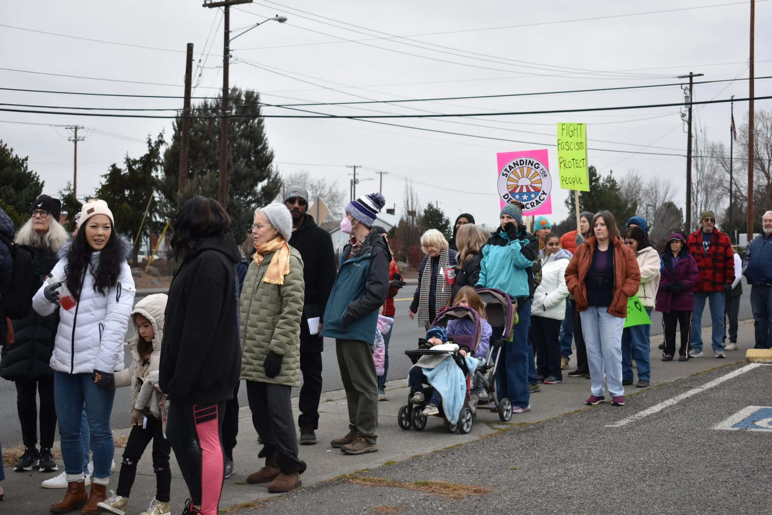 A group of diverse people participating in a protest or demonstration outdoors. Some are holding signs, one reading 'FIGHT Fascism. PROTECT people,' and another with a logo and the words 'STANDING FOR DEMOCRACY.' The weather appears cold and overcast, with many people wearing jackets, hats, and scarves.