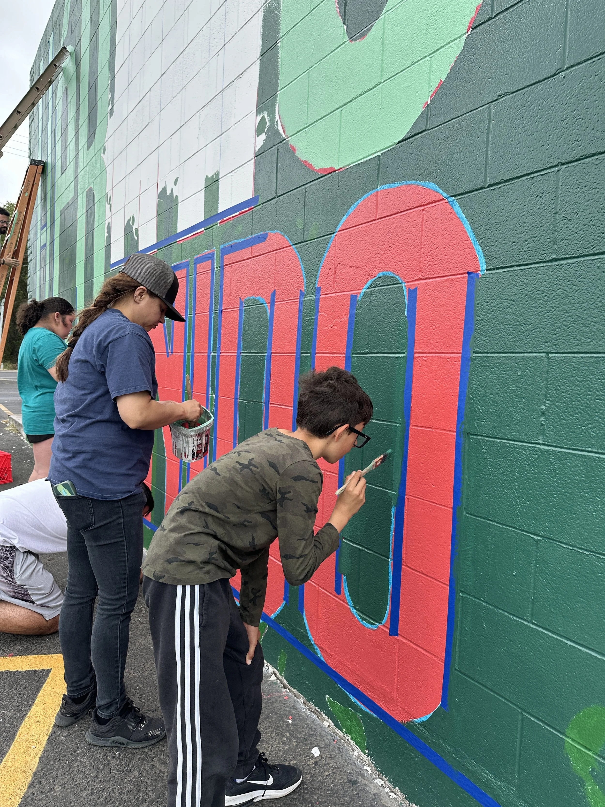 Young people painting a colorful mural on a brick wall outside in a parking lot.