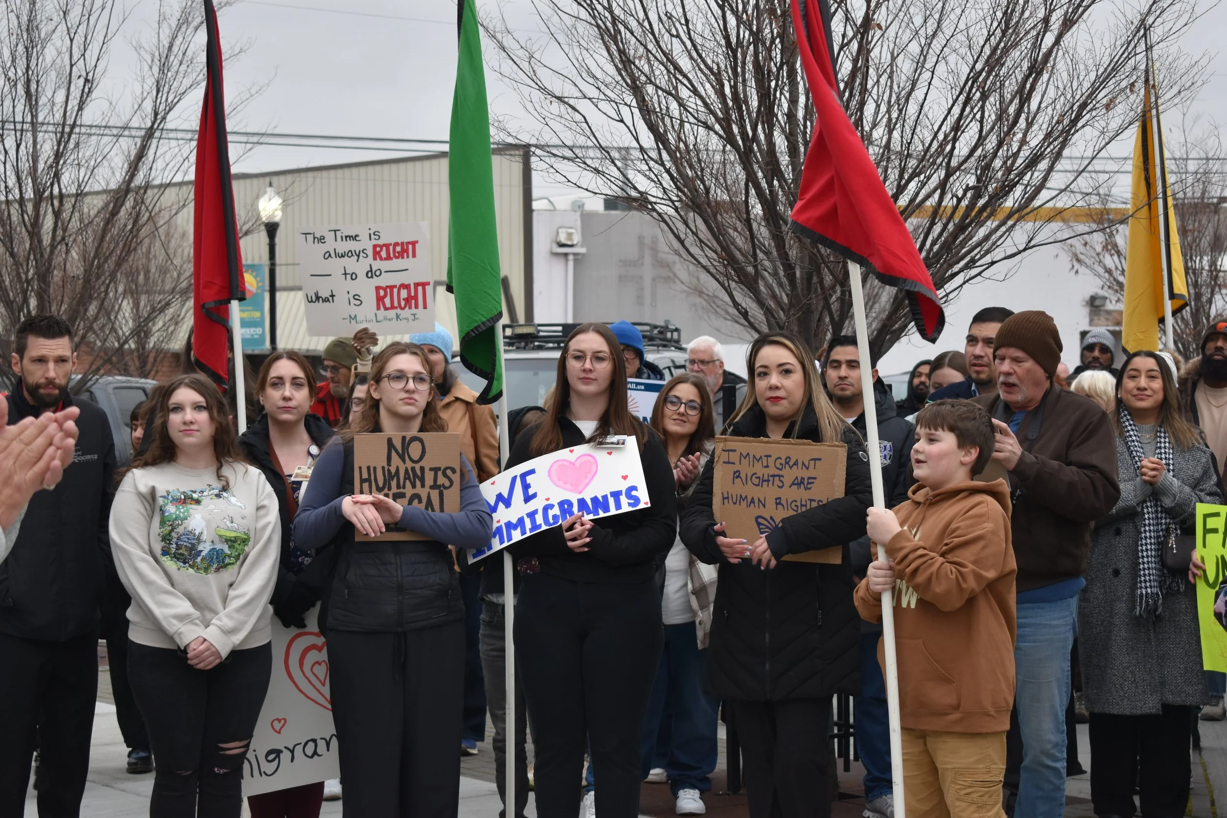 People protesting outdoors, holding signs about immigration rights and advocating for immigrants. Some signs include messages like 'The Time is always RIGHT to do what is RIGHT,' 'NO HUMANS IS ILLEGAL,' and 'IMMIGRANT RIGHTS ARE HUMAN RIGHTS.'