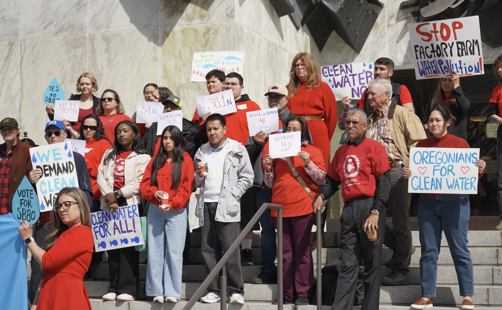 Group of people protesting with signs about clean water, pollution, and environmental issues, standing on steps outside a building.