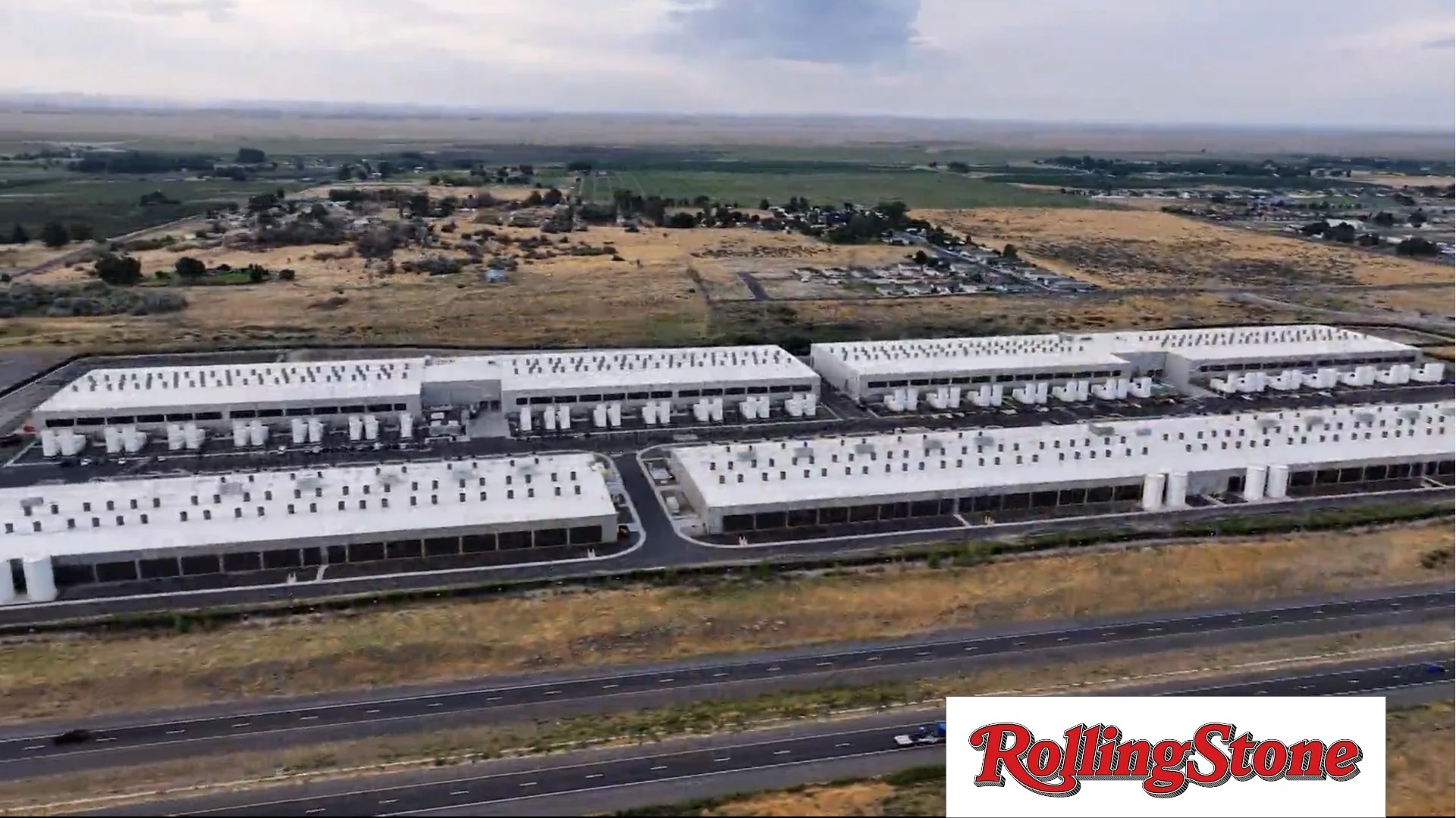 Aerial view of a large, modern building complex in a rural area on a cloudy day with fields and roads surrounding it, and the Rolling Stone logo in the bottom right corner.