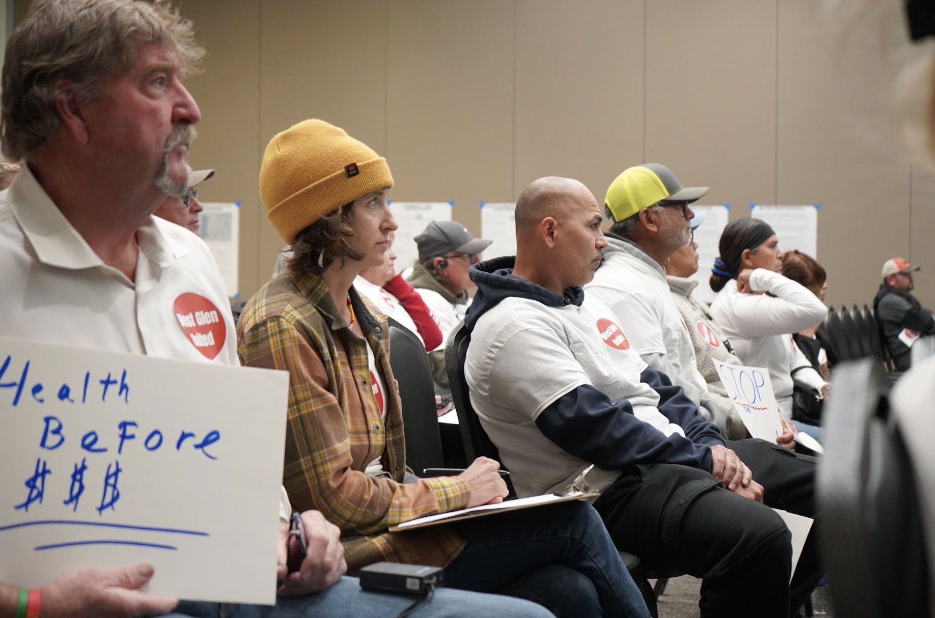 A diverse group of people sitting in a conference room, attending a protest or rally. Some are wearing white shirts with red heart stickers that say 'Health & Bmore,' and others are holding signs and listening attentively.
