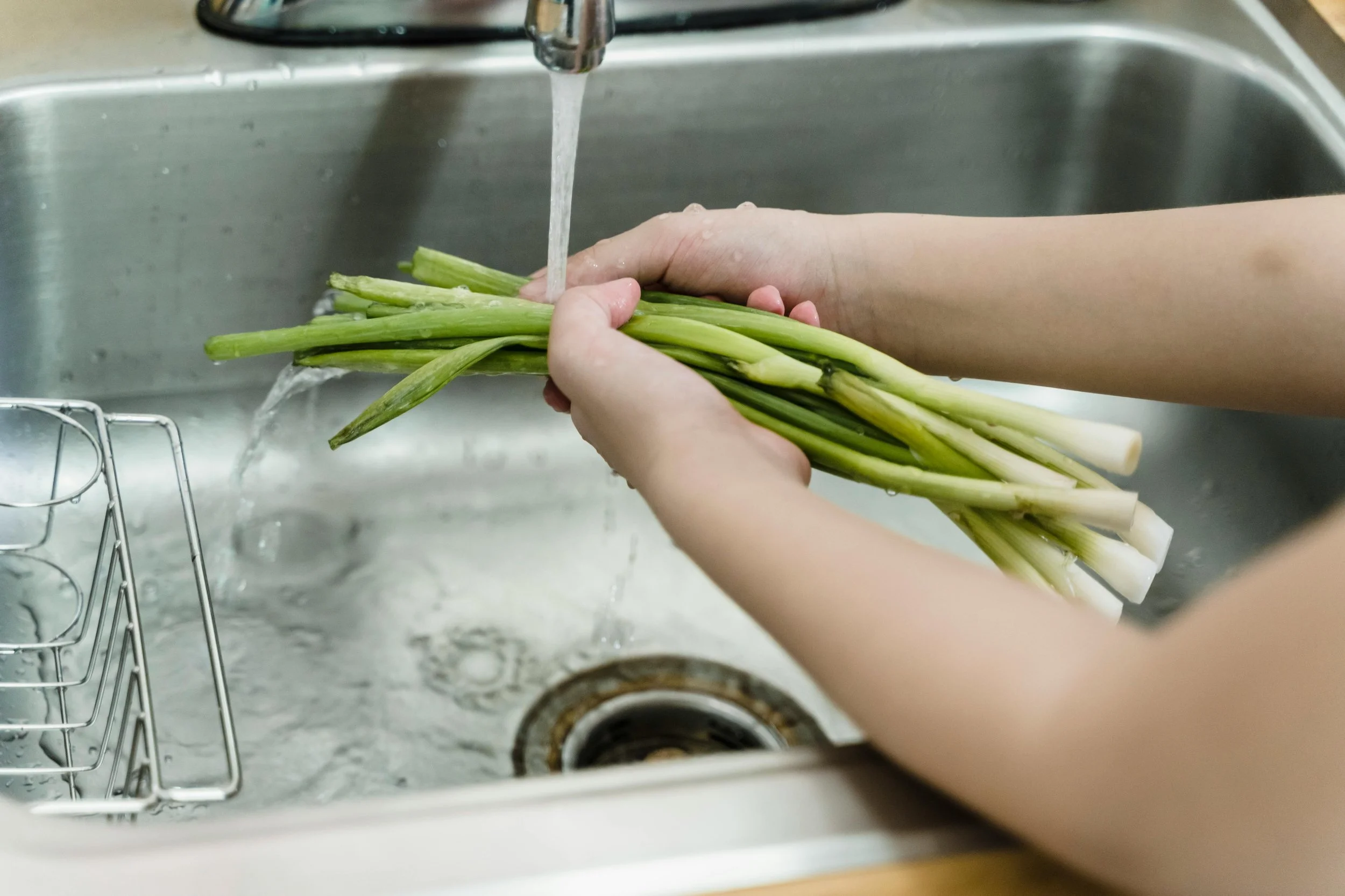 Person washing fresh green onions in a kitchen sink with running water.