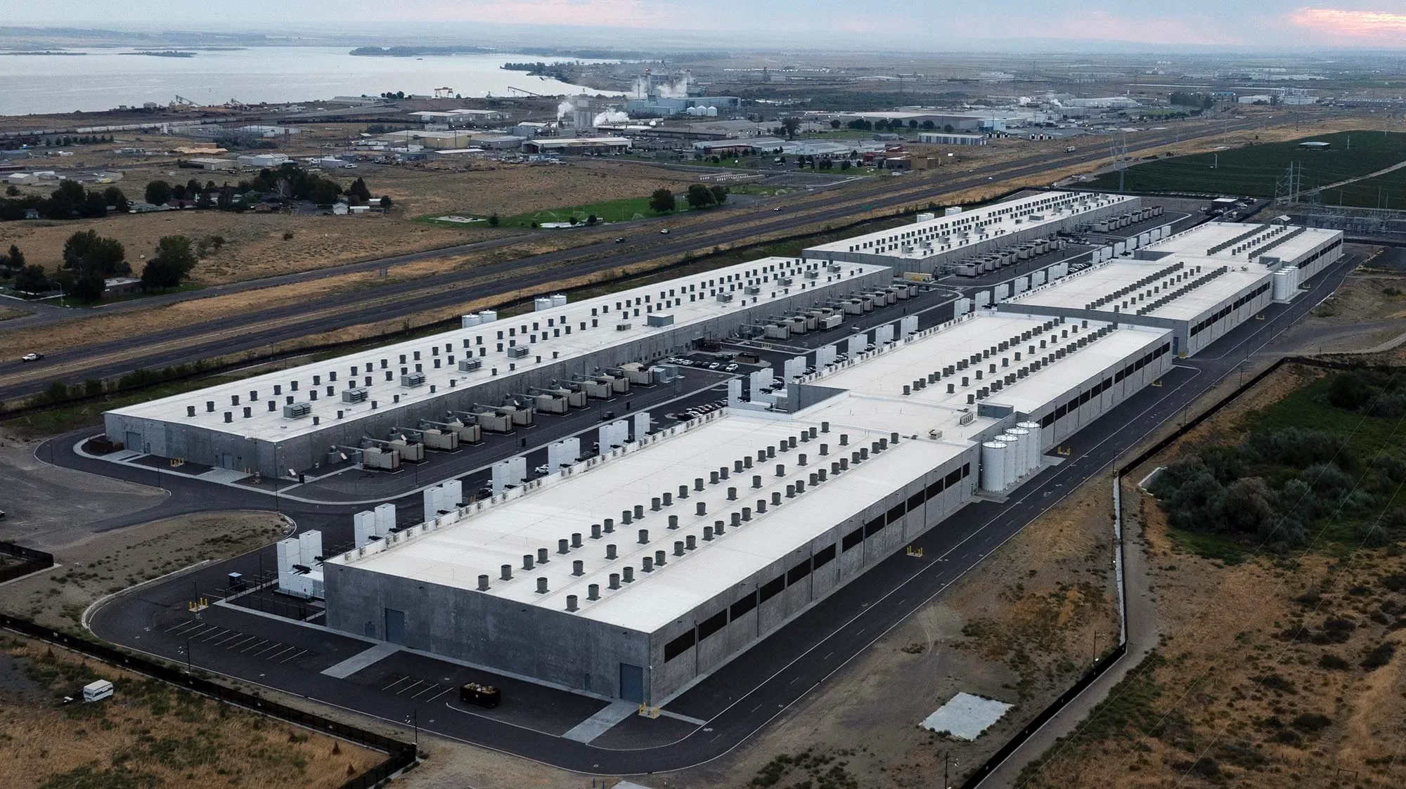 Aerial view of a large industrial building with white roof, multiple HVAC units, and parking lot, with fields, roads, and water in the background.