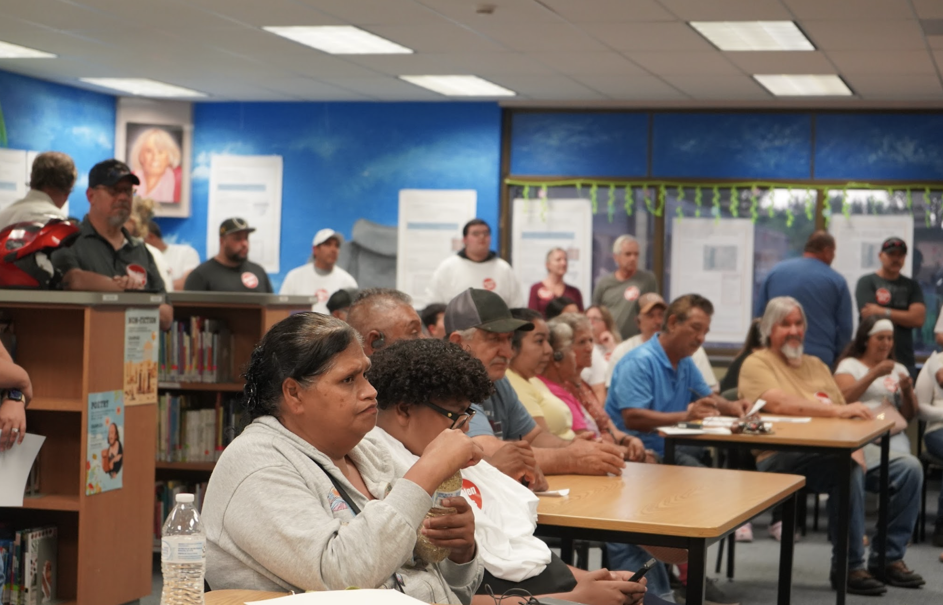 A large group of diverse people sitting in rows at tables in what appears to be a community hall or library, attending a event or meeting. Most are focused on something out of the frame, with some looking at their phones. The room has a colorful mural and framed posters on the walls, a bookshelf at the front, and the ceiling has fluorescent lights.