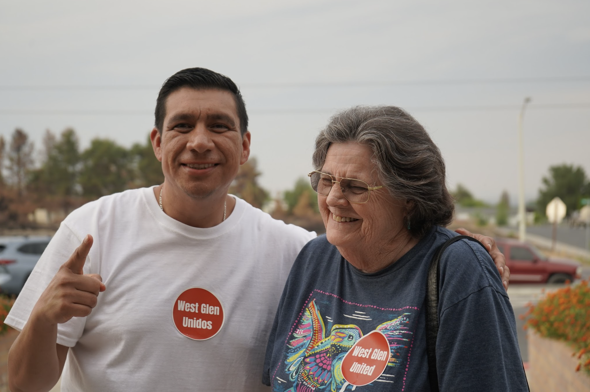 A young man with short dark hair and wearing a white t-shirt with a red sticker reading 'West Glen Unidos' is smiling and pointing at the camera. An older woman with short gray hair, glasses, and wearing a dark t-shirt with colorful designs, is smiling and looking at the young man. They are outdoors with trees and parked cars in the background.
