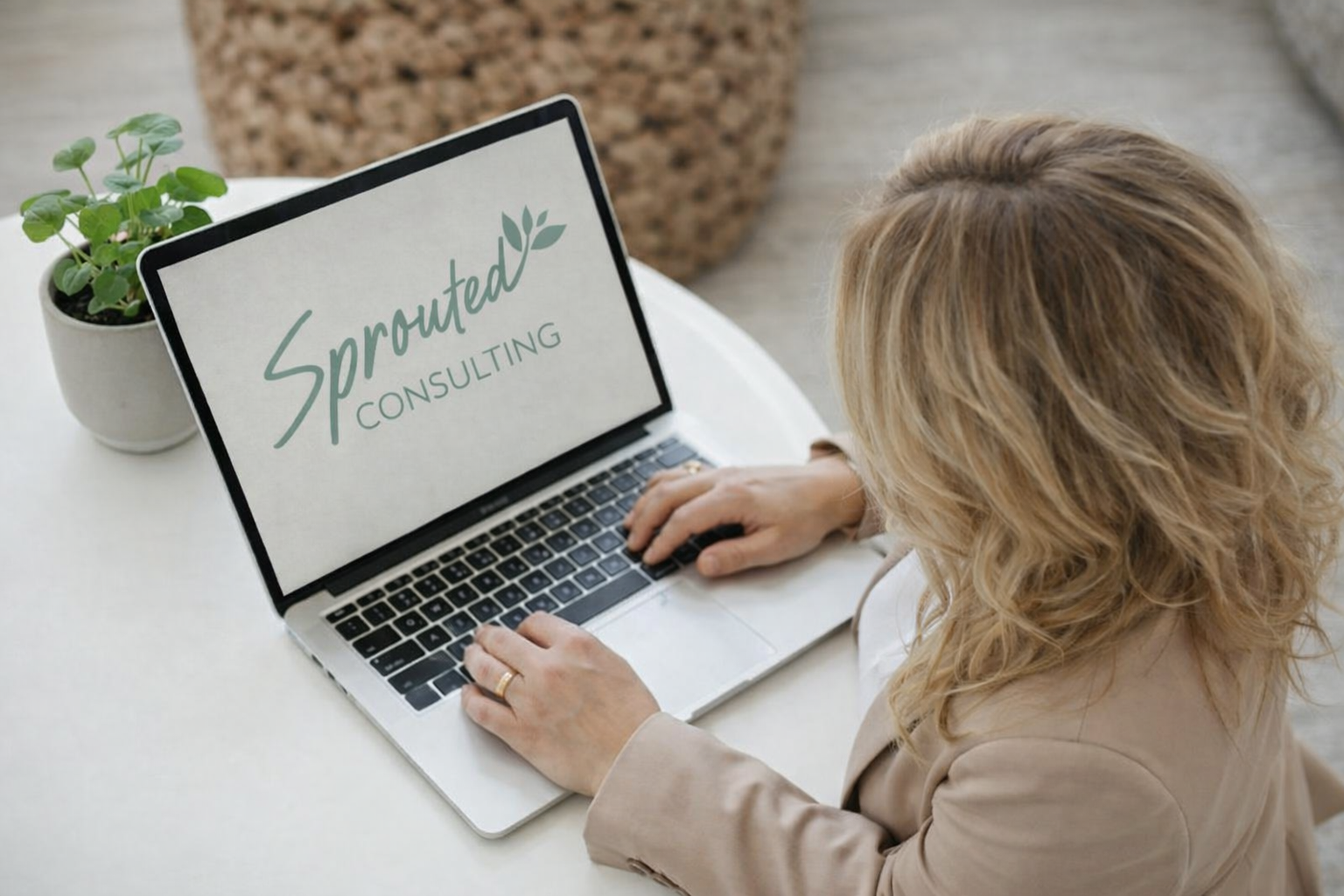 Cara with blonde curly hair working on a laptop at a white desk. The laptop screen shows the text 'Sprouted Consulting' with a leaf graphic. There is a small potted plant on the desk.