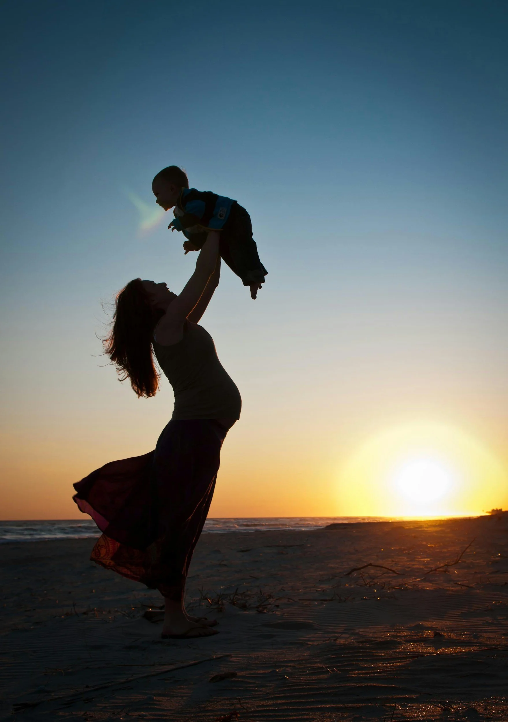 A silhouette of a woman lifting a child in the air on a beach at sunset.
