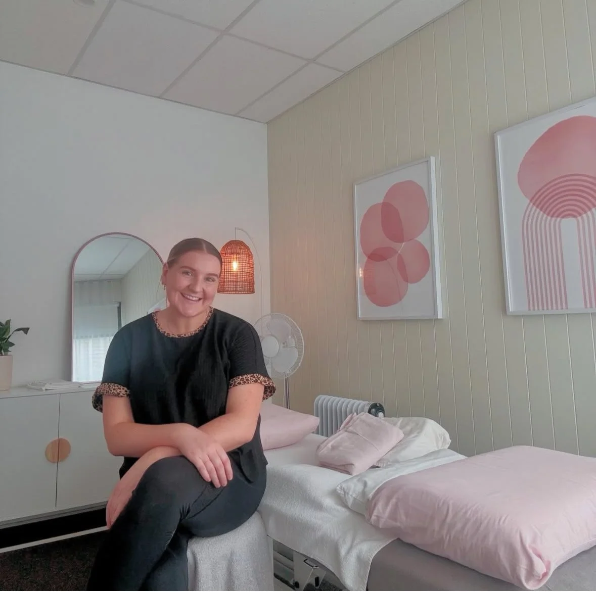 A woman sitting on a stool in a bedroom, smiling at the camera, with two abstract pink artwork pieces on the wall, a mirror, a lamp, a fan, a radiator, and a bed with pillows and a pink blanket.