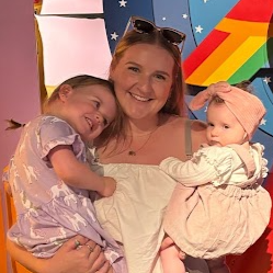 A woman holding two young girls, one on each side. The girl on the left is smiling and resting her head on the woman's shoulder, wearing a purple dress. The girl on the right is looking to the side, wearing a pink dress and a pink hat. The background features colorful rainbow and star decorations.