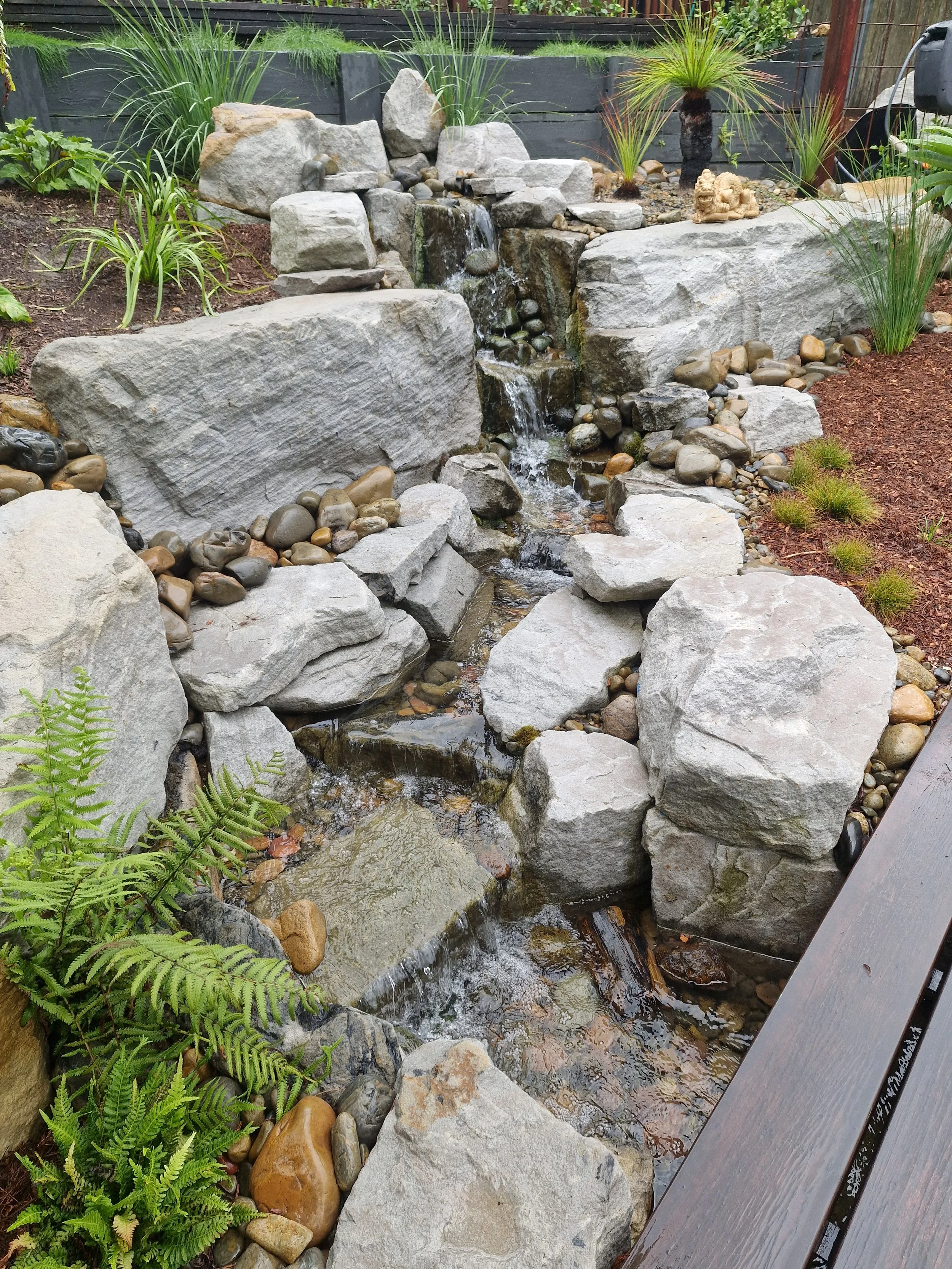 Small waterfall flowing over rocks in a landscaped garden with various plants, pebbles, and a wooden bench nearby.