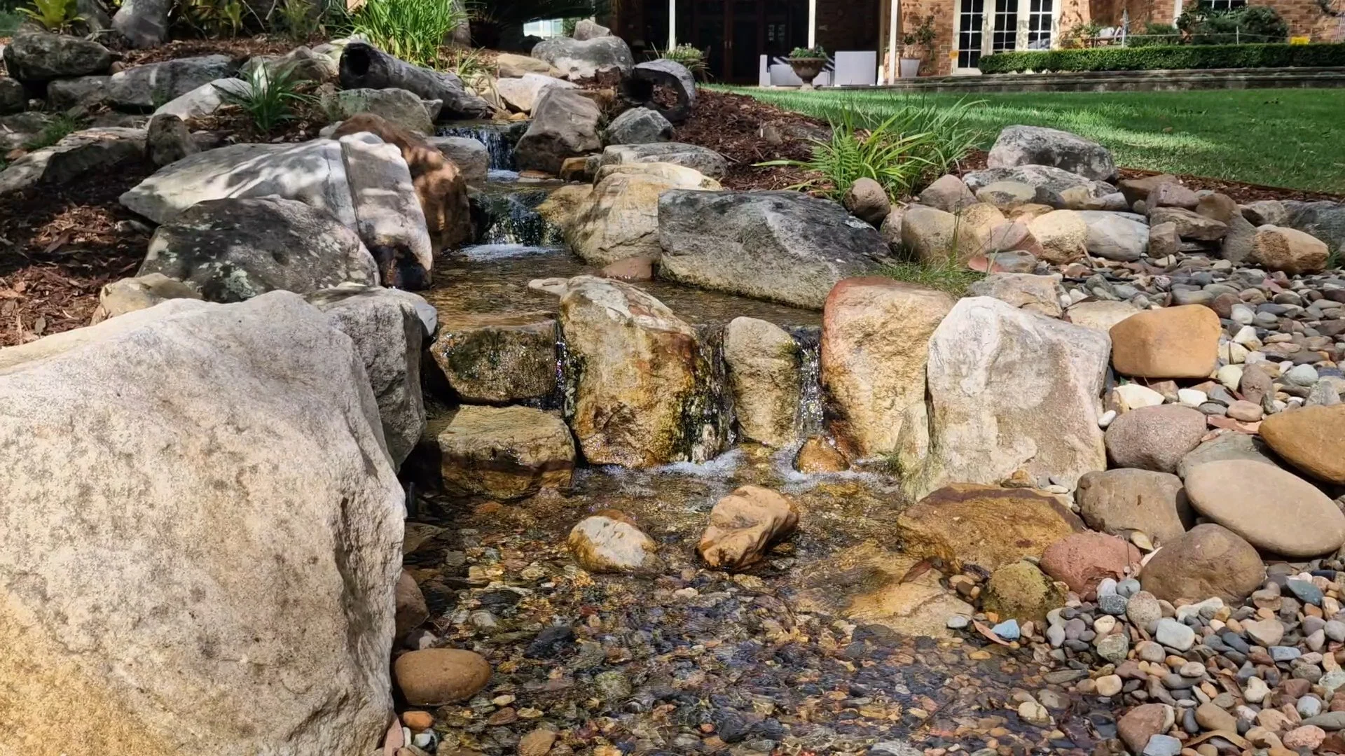 A backyard landscape with a small waterfall flowing over rocks, surrounded by various stones and a manicured lawn with shrubs and a brick house in the background.
