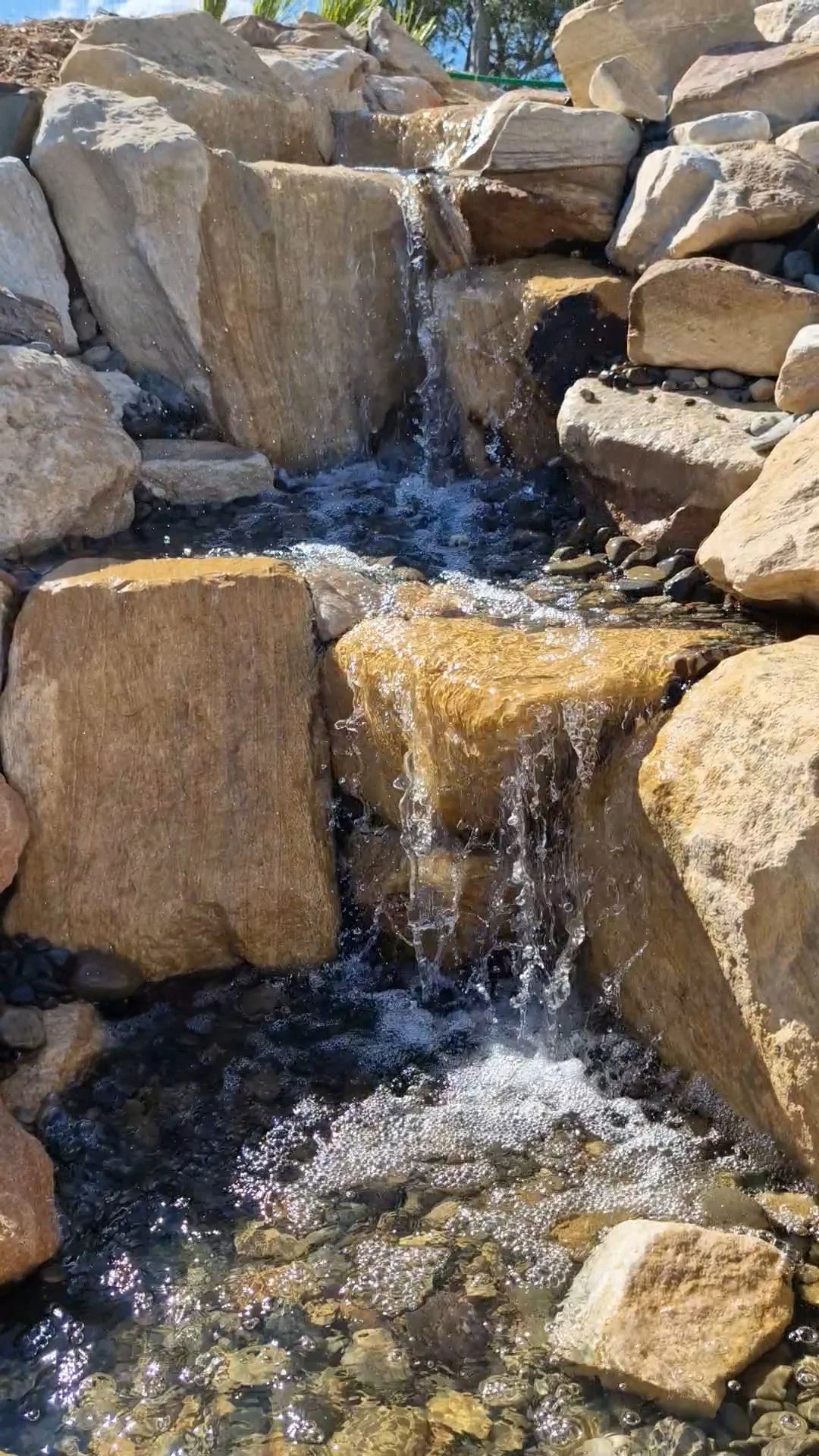 Small waterfall flowing over rocks in a rocky creek bed with clear water and sunlight reflections.