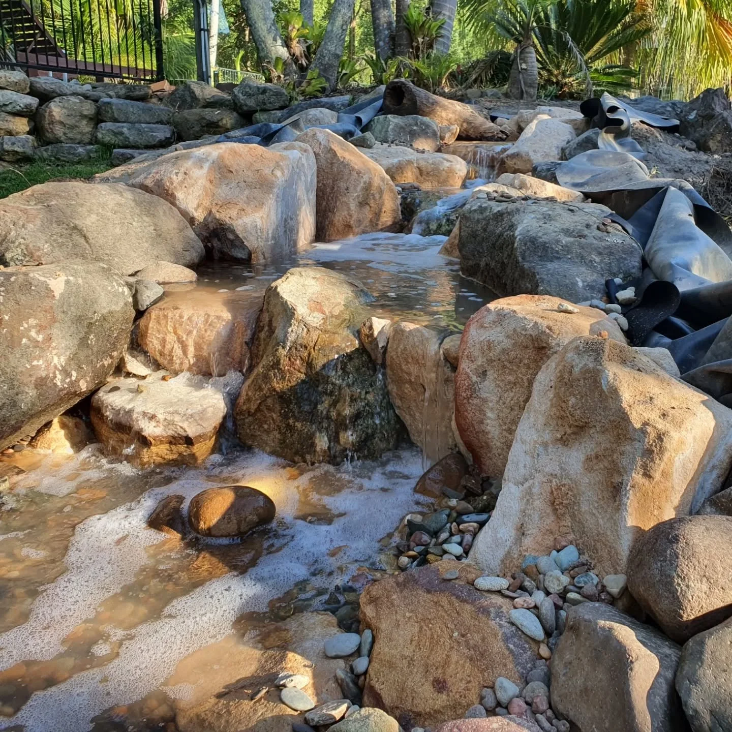 A small man-made stream with large rocks and pebbles, surrounded by greenery and palm trees, with some plastic sheeting extending over the rocks.